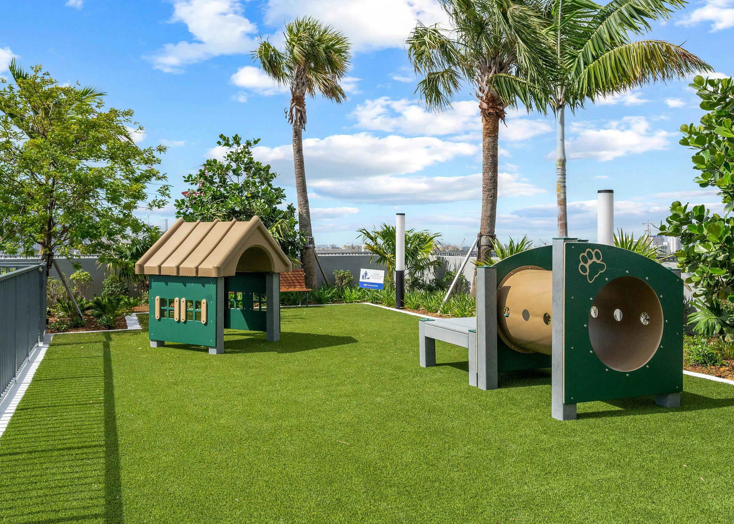 A daytime view of the rooftop gated pet park at AMLI Wynwood apartments surrounded by trees with green turf and two play tunnels