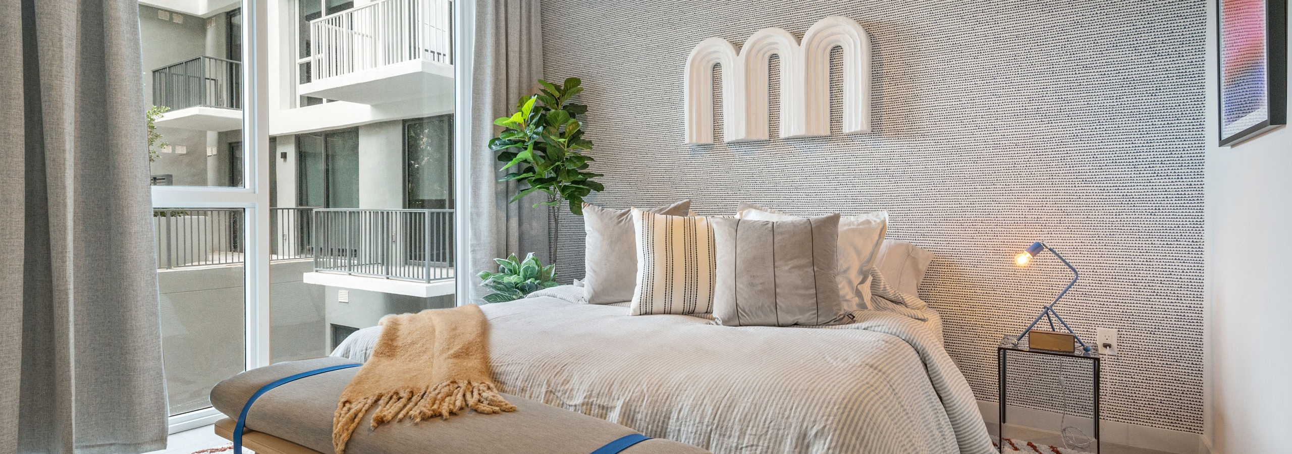 A bedroom at AMLI Wynwood apartments with a textured grey accent wall showing mounted artwork and a queen sized bed with modern furnishings