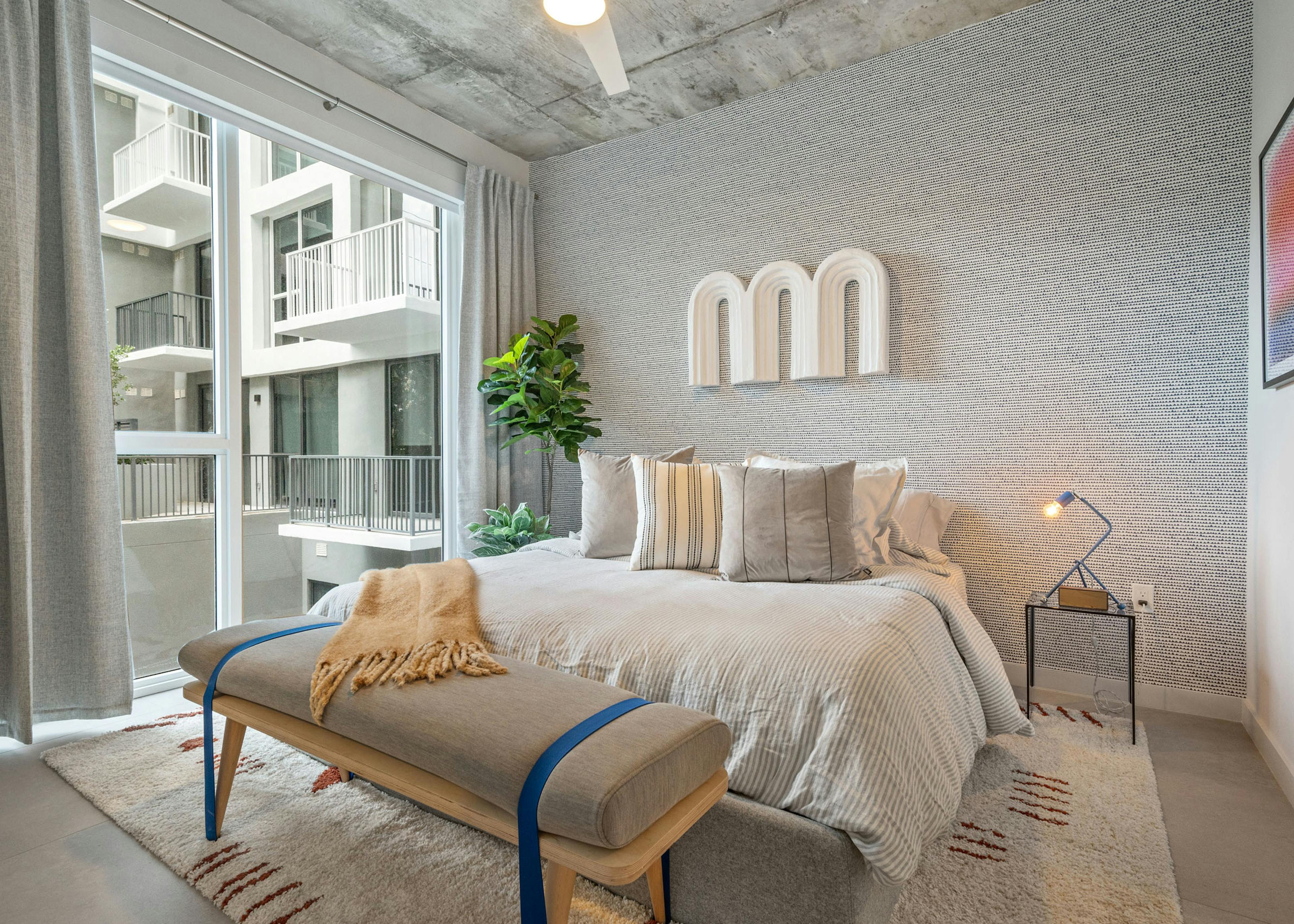 A bedroom at AMLI Wynwood apartments with a textured grey accent wall showing mounted artwork and a queen sized bed with modern furnishings