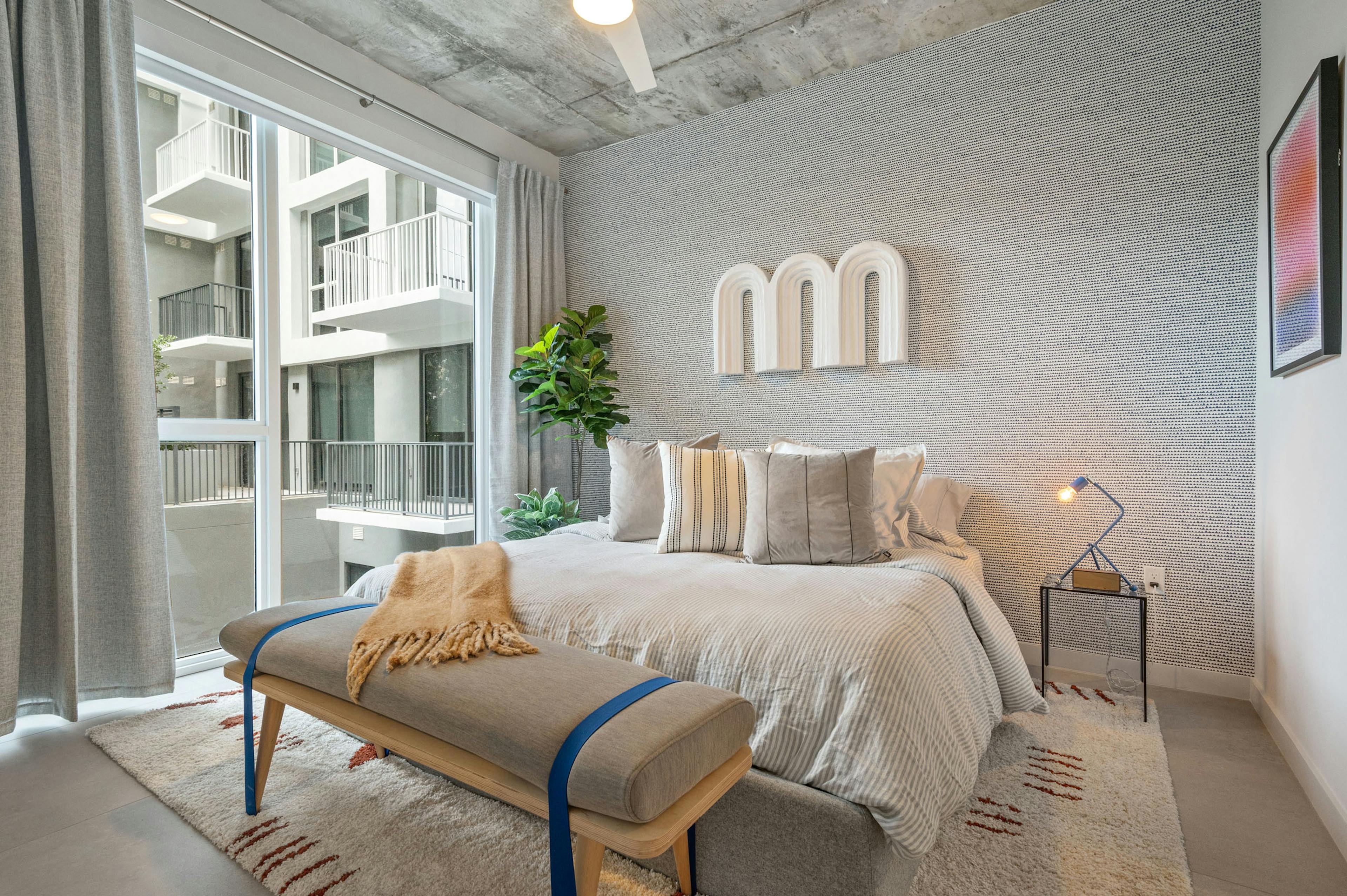 A bedroom at AMLI Wynwood apartments with a textured grey accent wall showing mounted artwork and a queen sized bed with modern furnishings