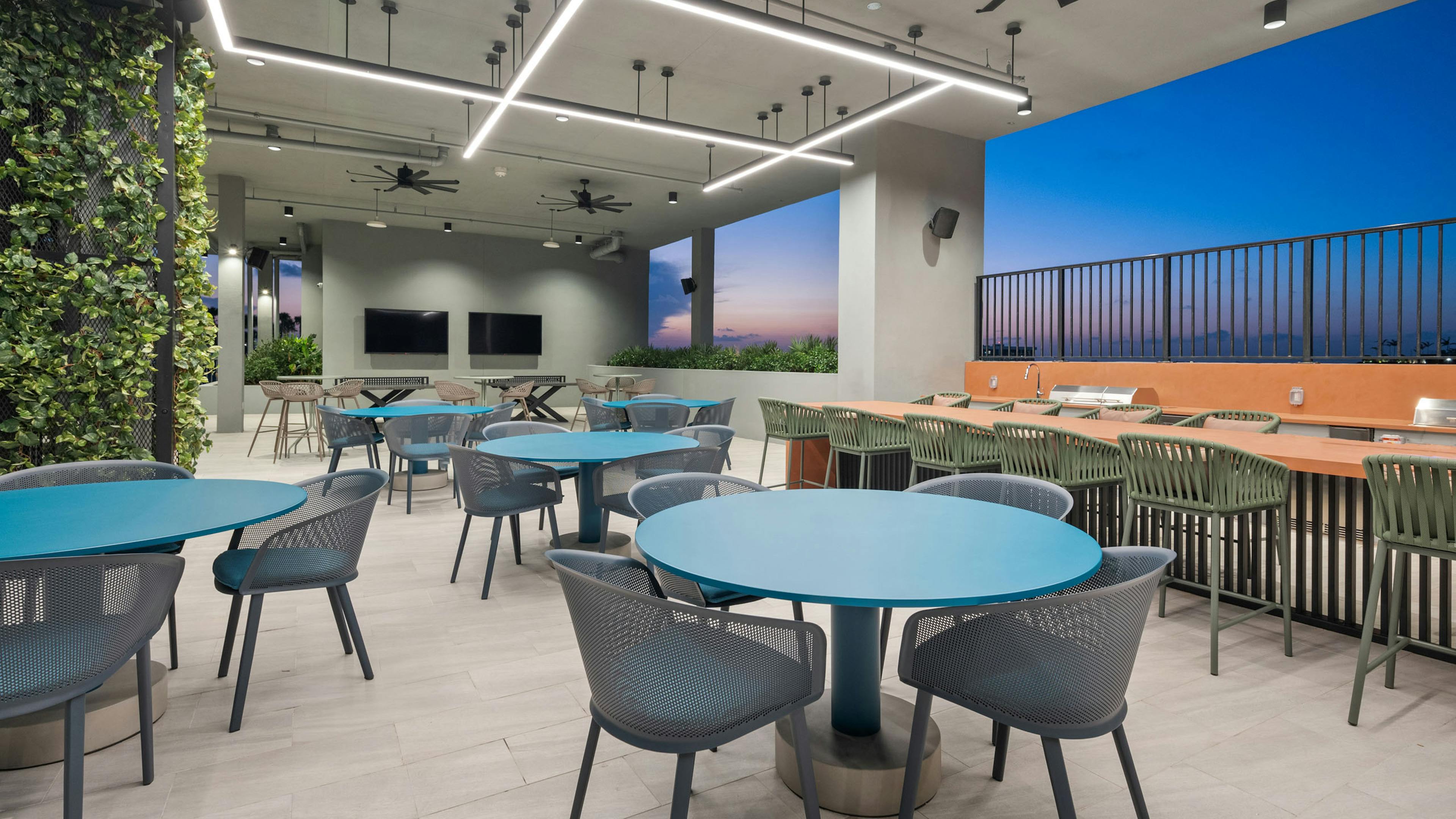 A covered rooftop space at AMLI Wynwood apartments with various sized tables and chairs and two grills overlooking a sunset view