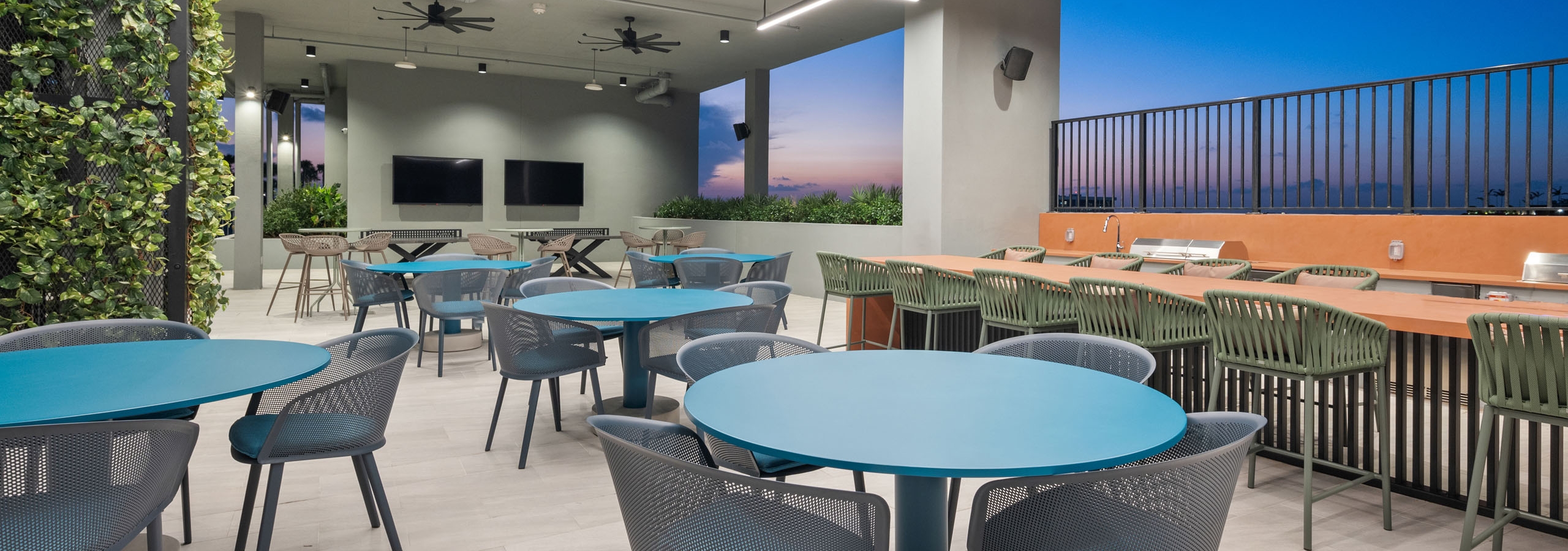 A covered rooftop space at AMLI Wynwood apartments with various sized tables and chairs and two grills overlooking a sunset view