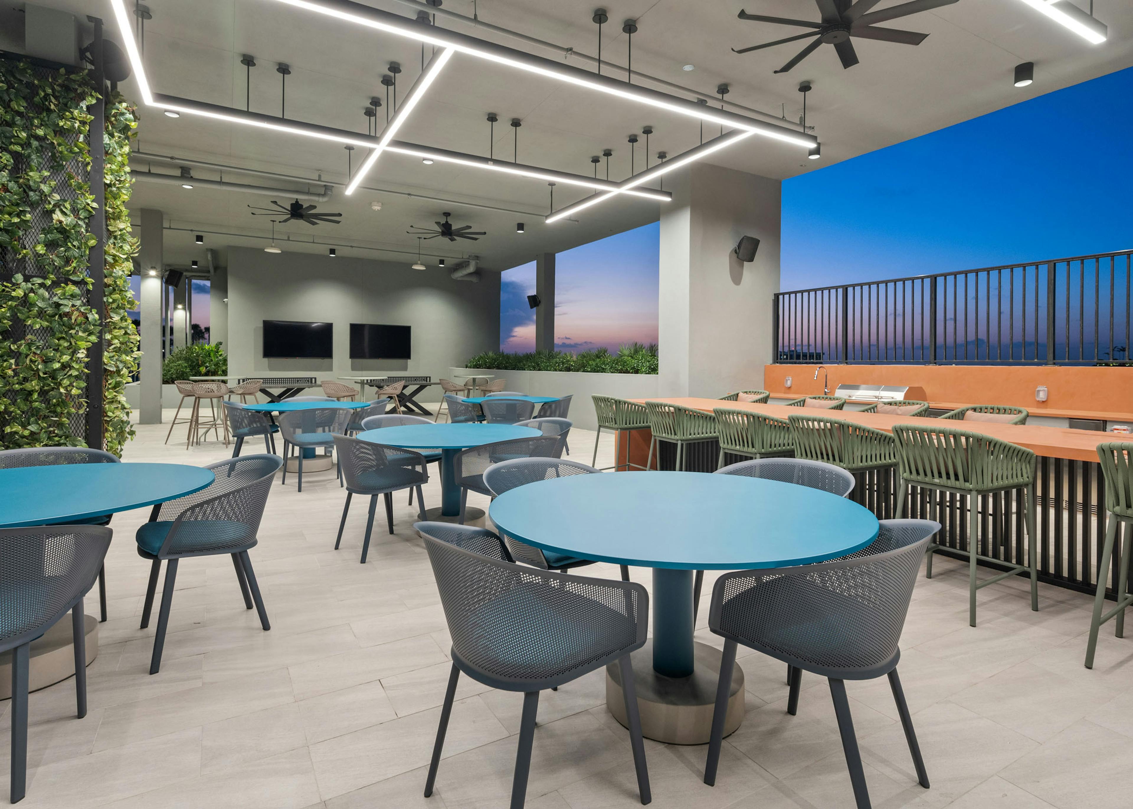 A covered rooftop space at AMLI Wynwood apartments with various sized tables and chairs and two grills overlooking a sunset view