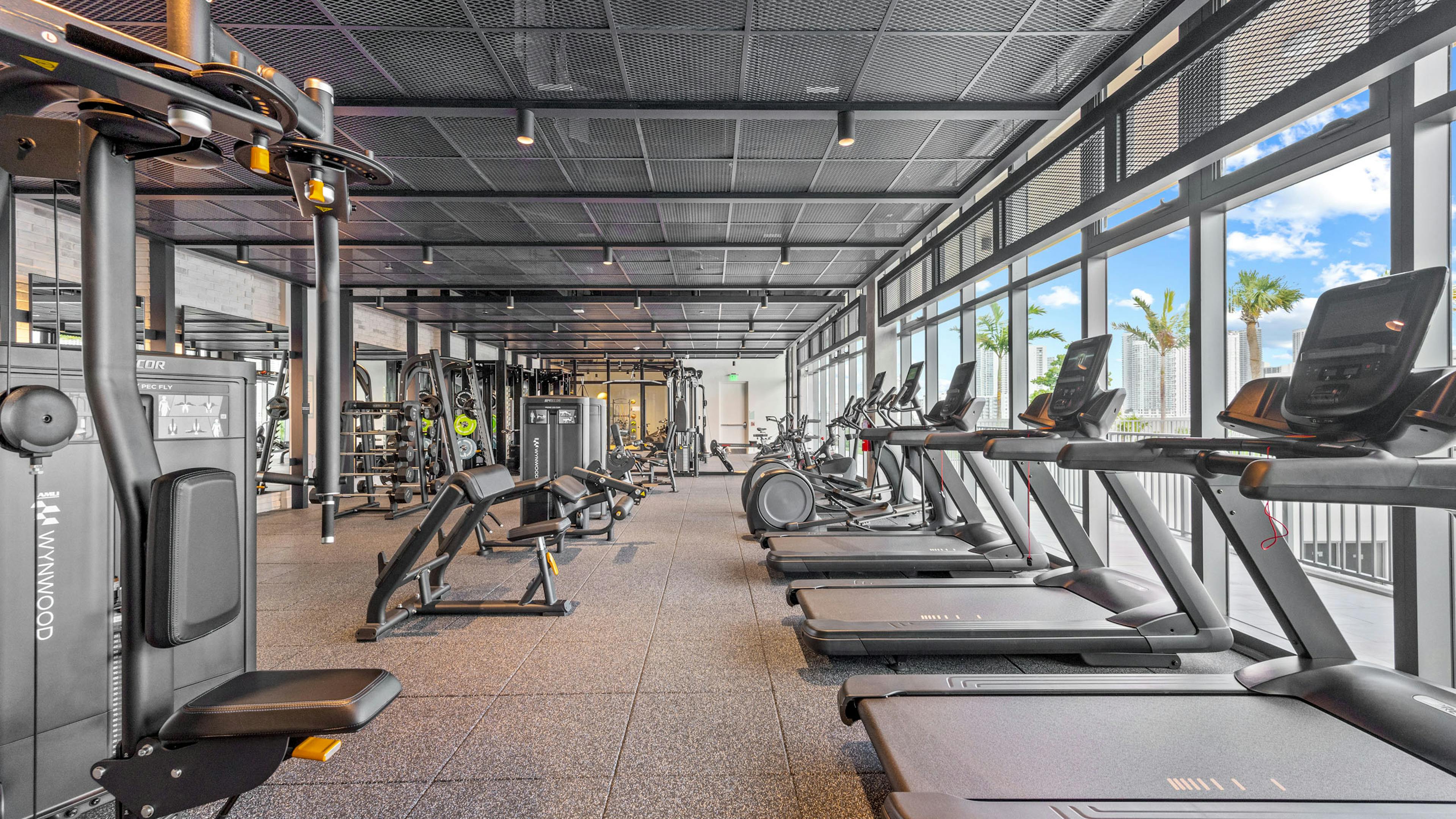 A large fitness center at AMLI Wynwood apartments featuring cardio and strength training equipment and windows with a view of palm trees