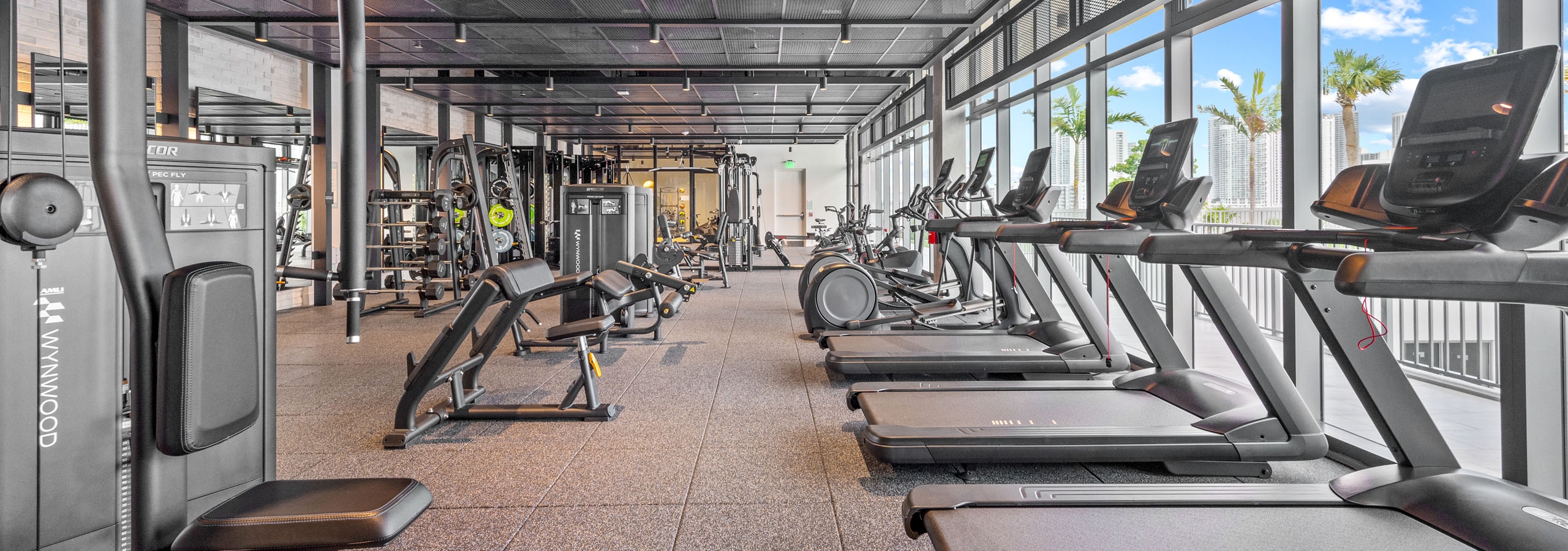 A large fitness center at AMLI Wynwood apartments featuring cardio and strength training equipment and windows with a view of palm trees