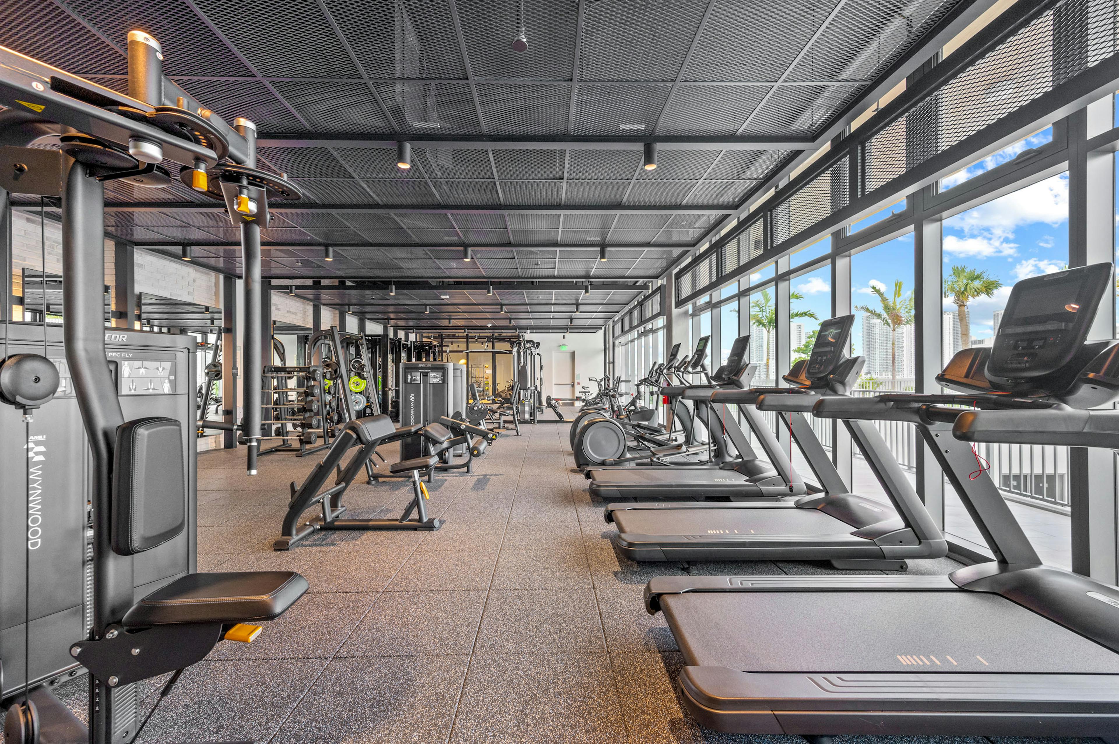 A large fitness center at AMLI Wynwood apartments featuring cardio and strength training equipment and windows with a view of palm trees