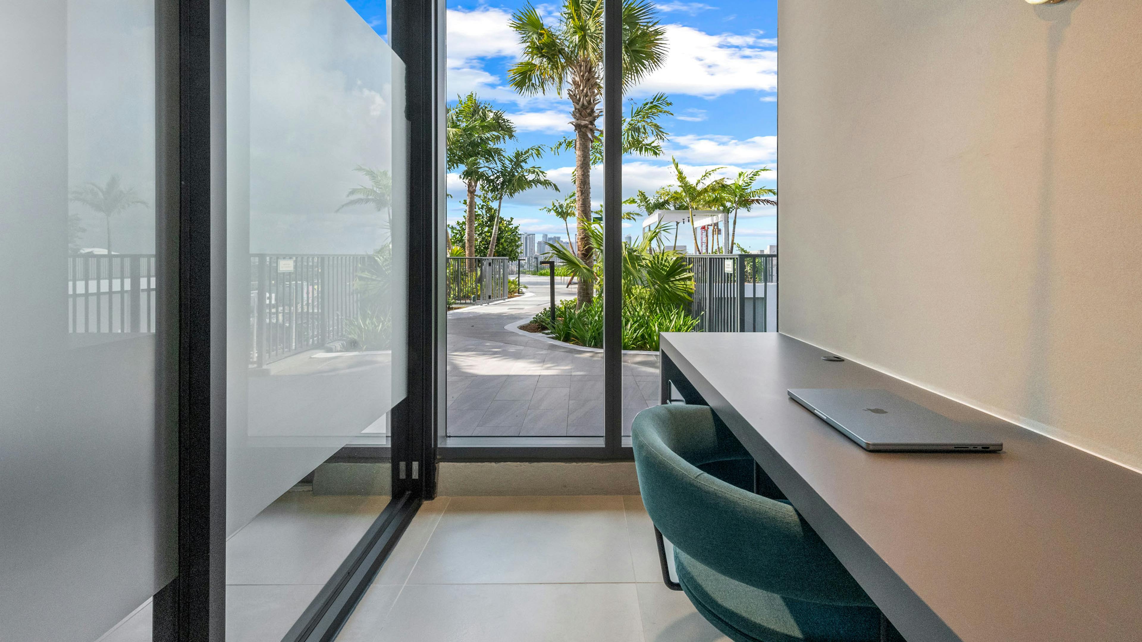 A glass office at AMLI Wynwood apartments with a green chair and laptop on a desk looking onto a sunny rooftop path with palm trees