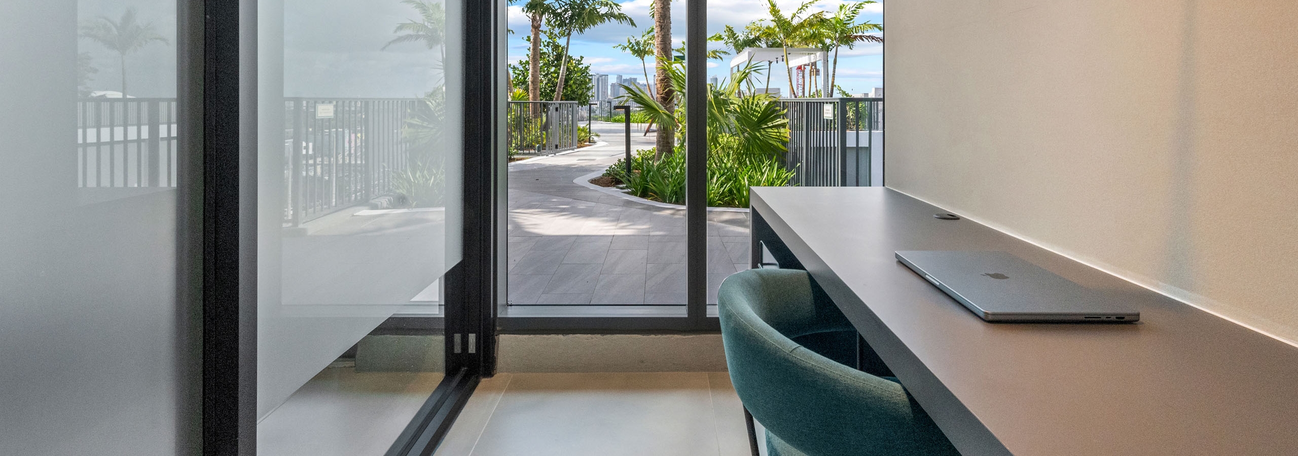 A glass office at AMLI Wynwood apartments with a green chair and laptop on a desk looking onto a sunny rooftop path with palm trees