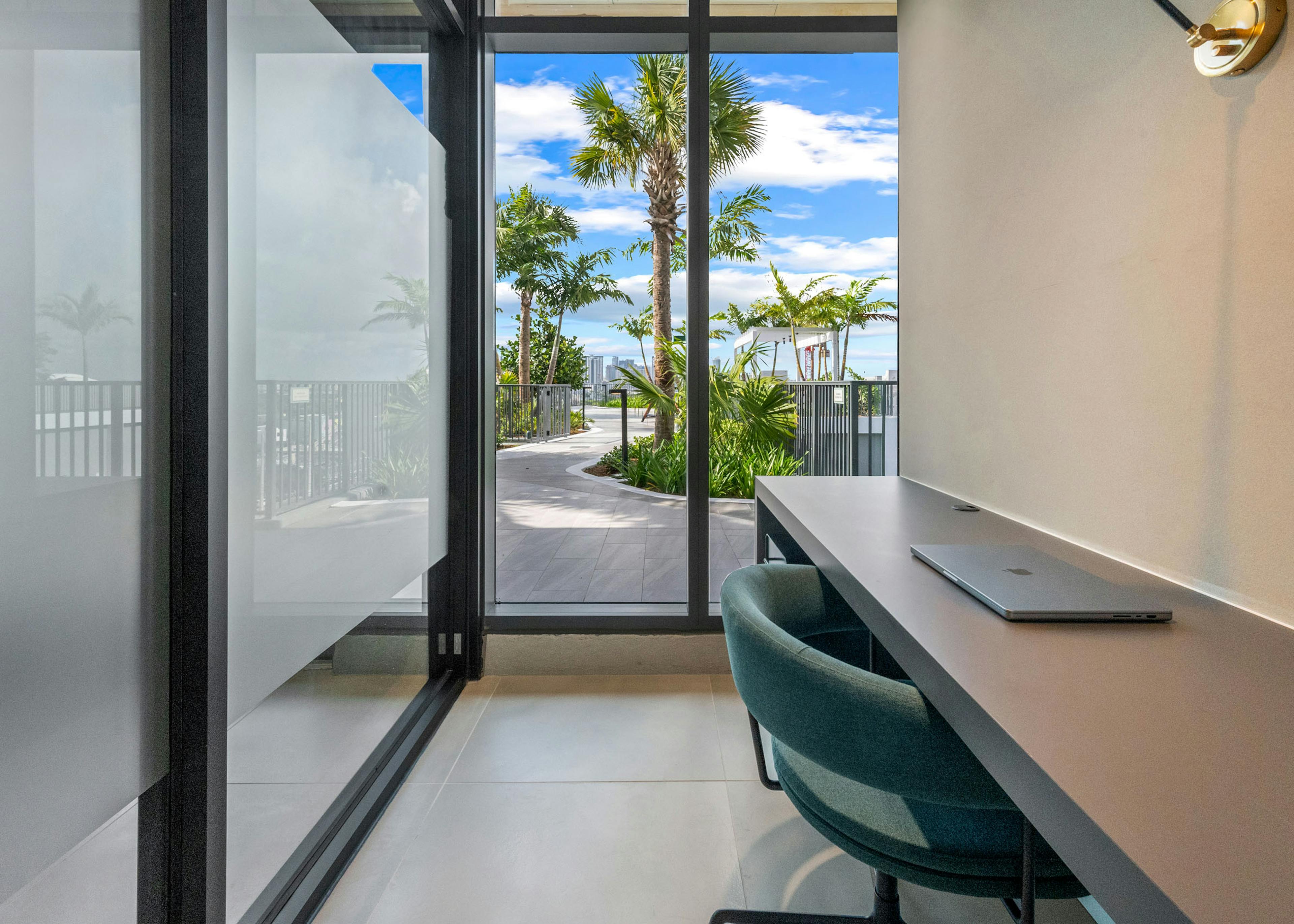 A glass office at AMLI Wynwood apartments with a green chair and laptop on a desk looking onto a sunny rooftop path with palm trees