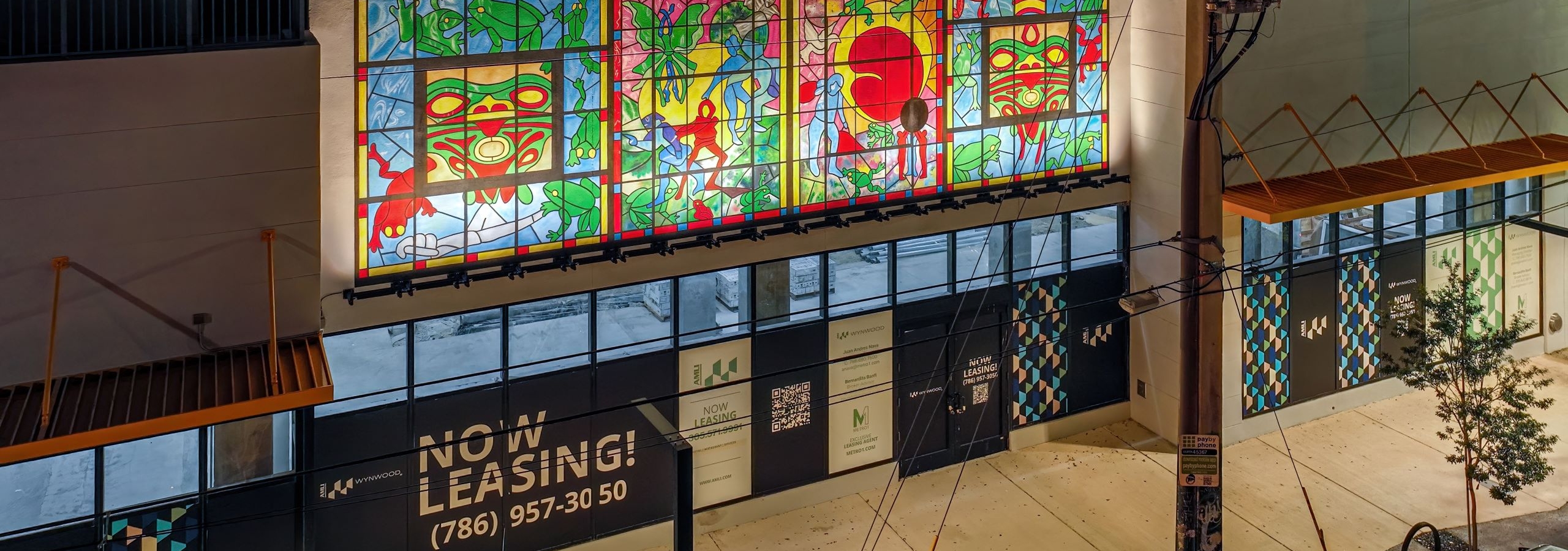 AMLI Wynwood retail space at night with an colorful mural above NOW LEASING windows on a tree lined street