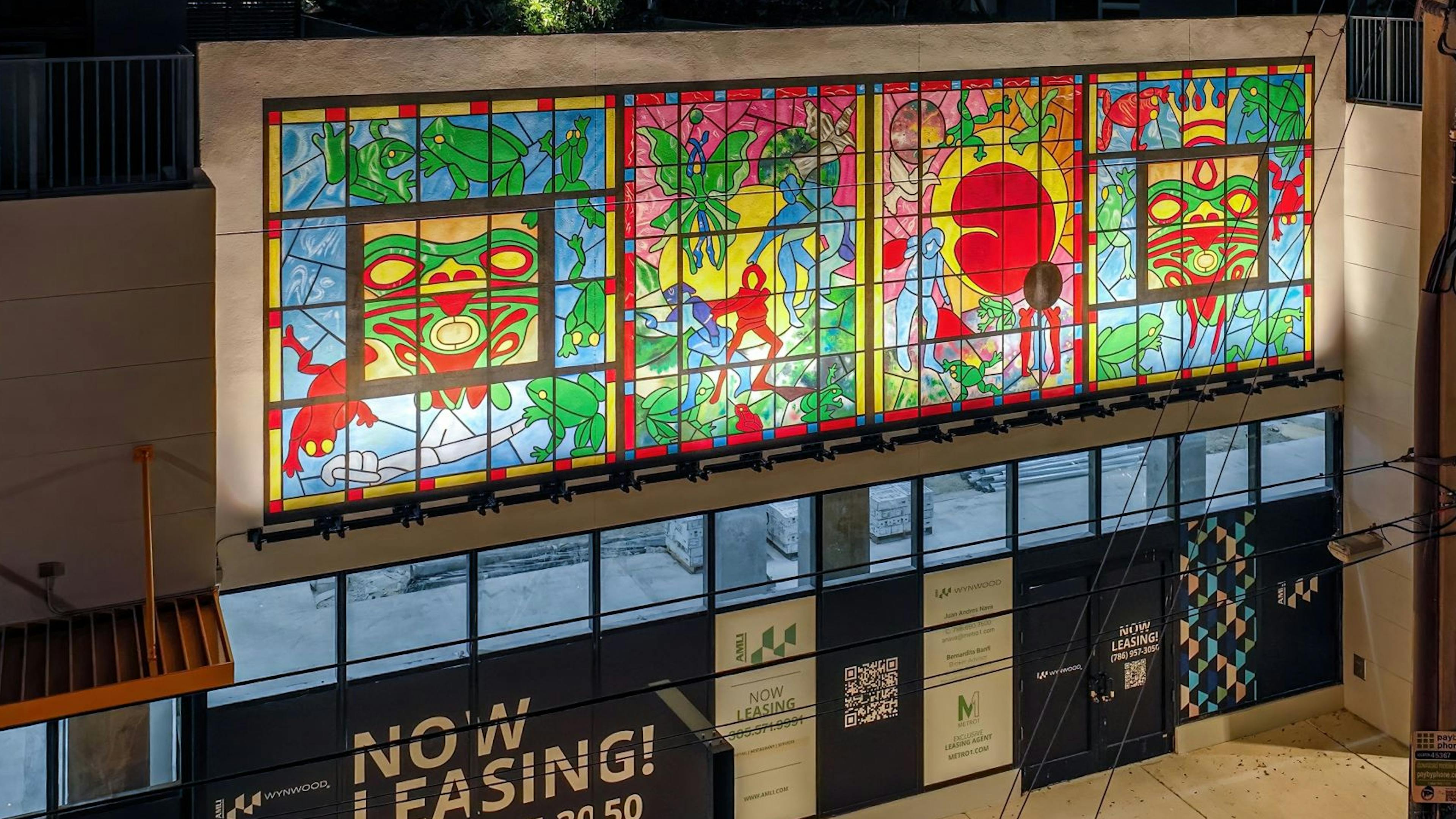 AMLI Wynwood retail space at night with an colorful mural above NOW LEASING windows on a tree lined street