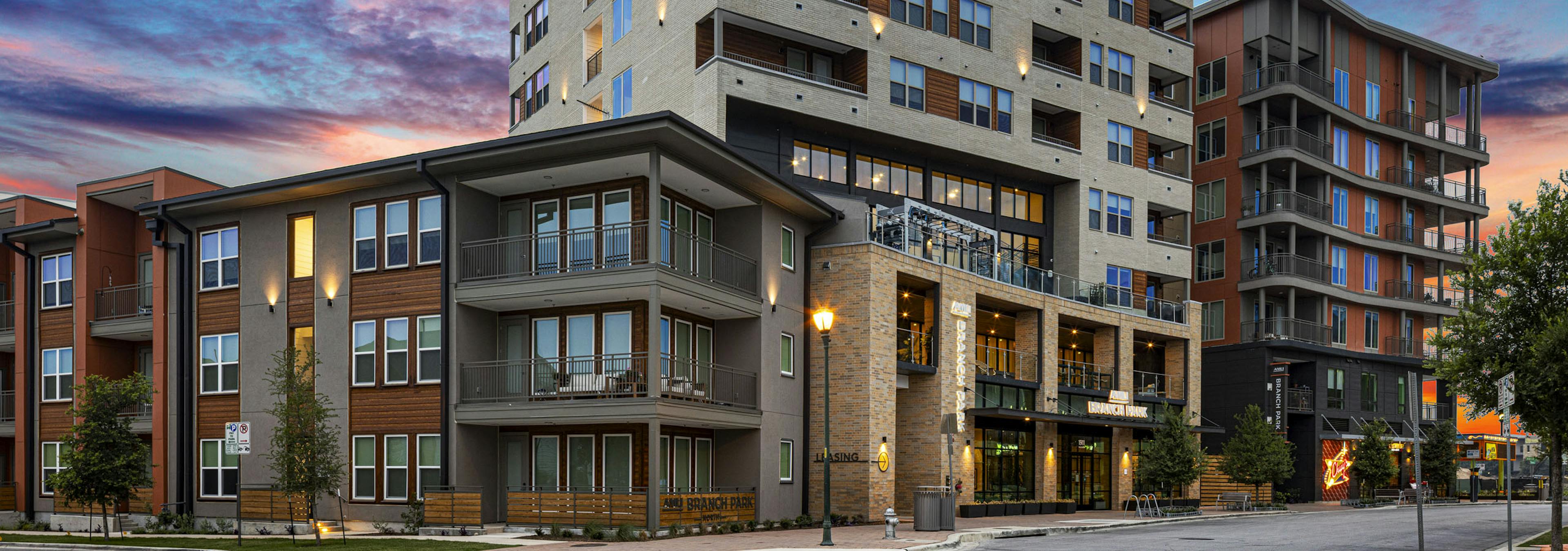 AMLI Branch Park multi-story tan and rust colored wood and brick apartment building with balconies on tree lined street at dusk.

