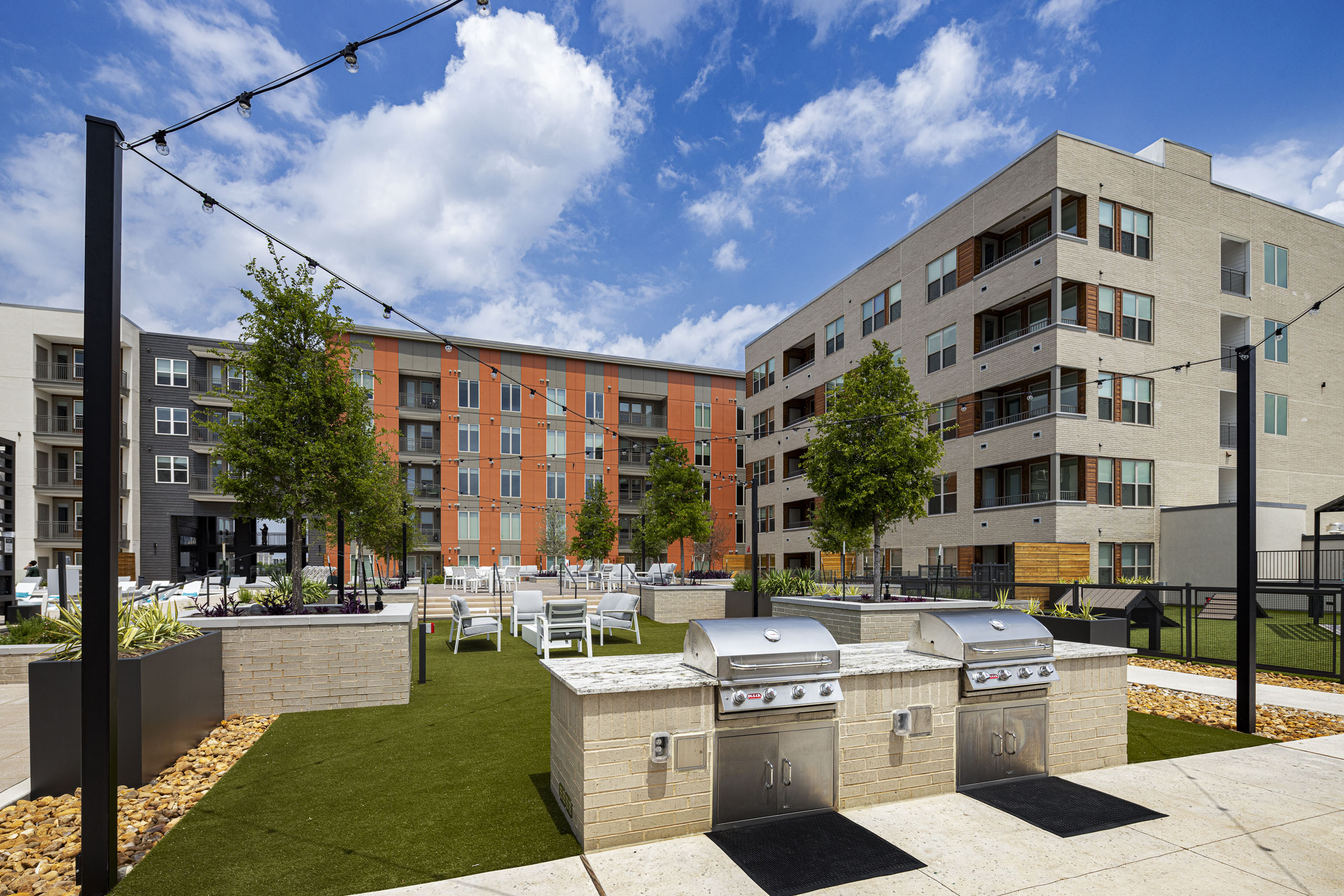 Outdoor community area at AMLI Branch Park with two BBQ grills on a stone platform, surrounded by green turf and string lights overhead.