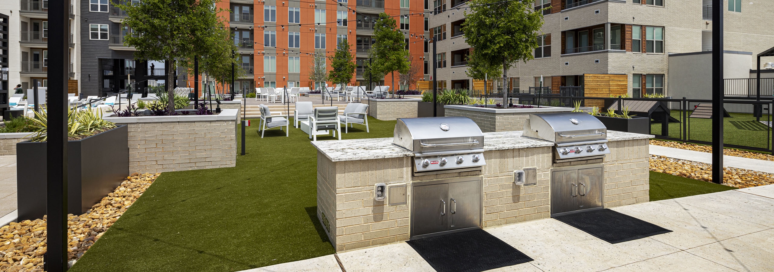 Outdoor community area at AMLI Branch Park with two BBQ grills on a stone platform, surrounded by green turf and string lights overhead.