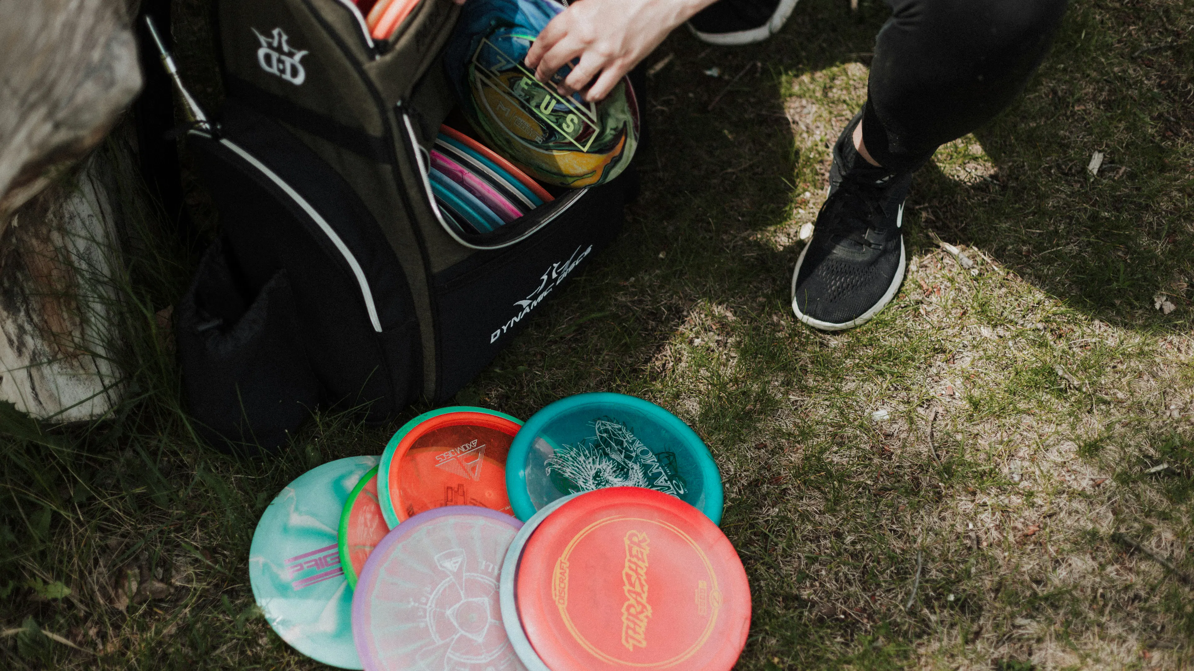 A person crouched on the grassy ground pulling disc golf discs out of a big bag