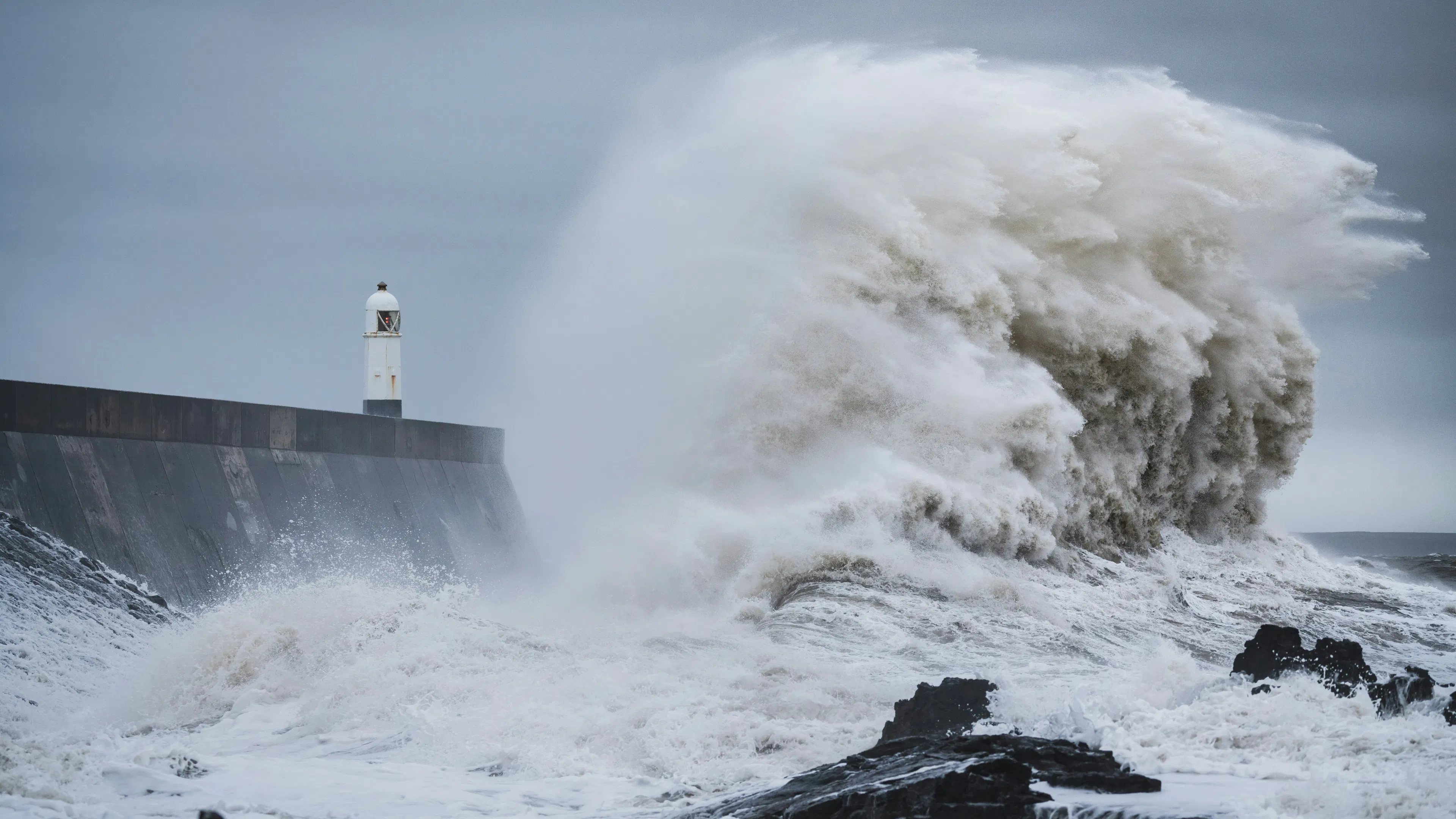 A large wave of dark stormy water crashing onto a breaker with a small lighthouse on a dark stormy day