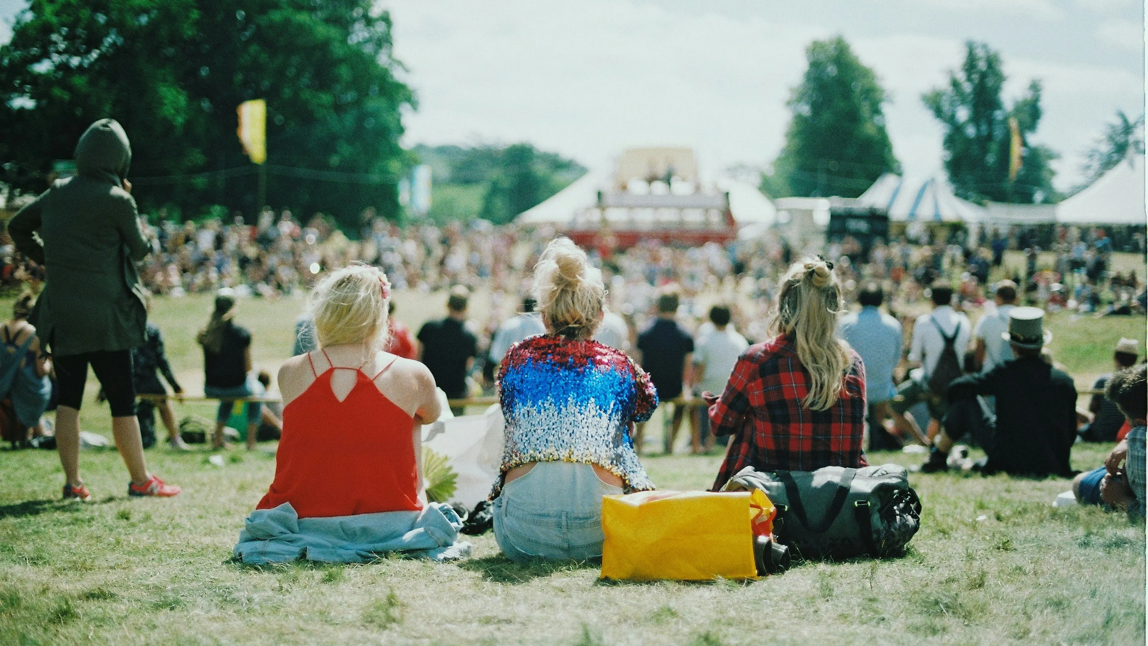 Three people sitting on a picnic blanket on a grassy lawn facing a music stage