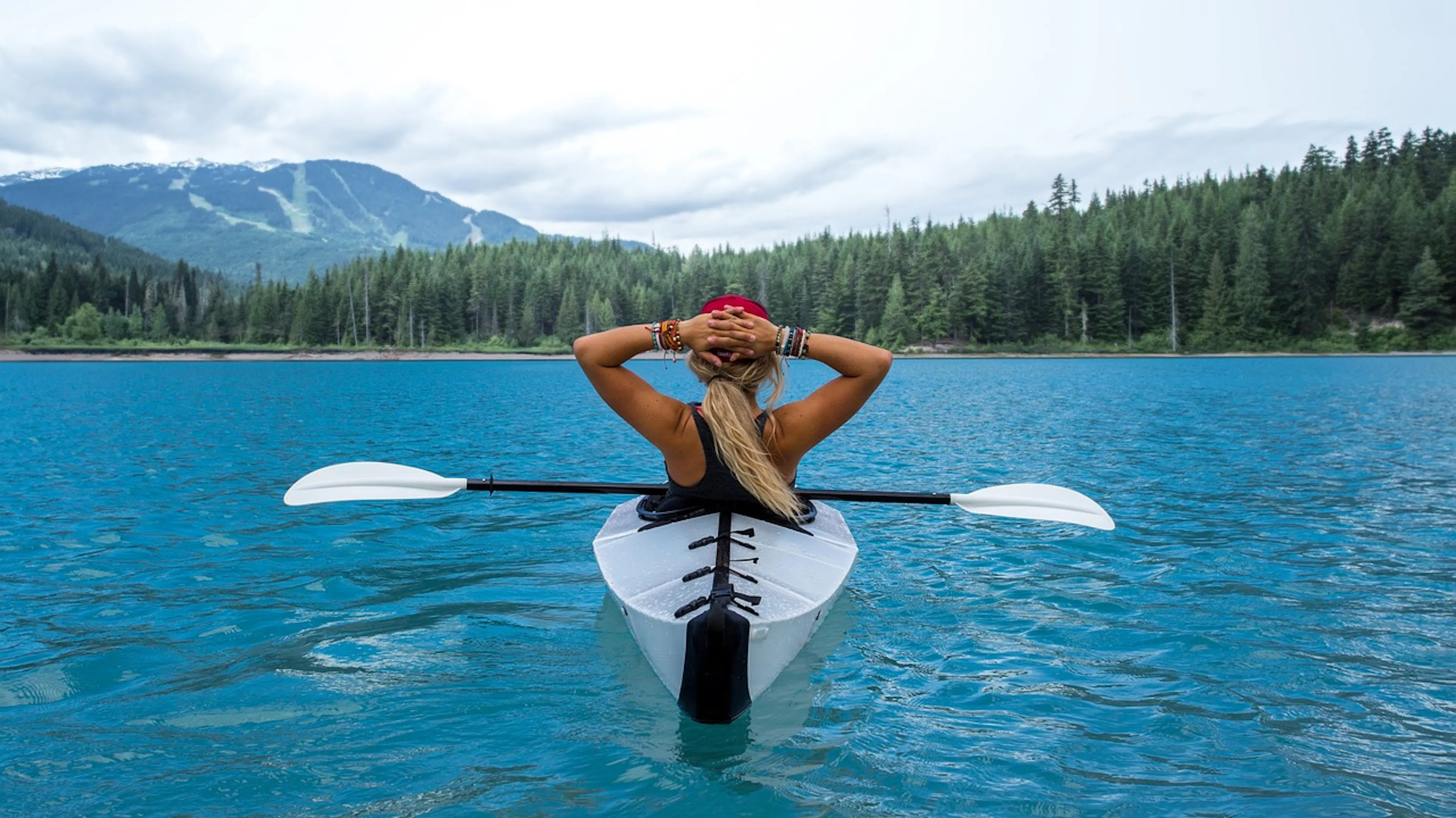 woman in white kayak with paddle in blue water in front of mountains with green trees