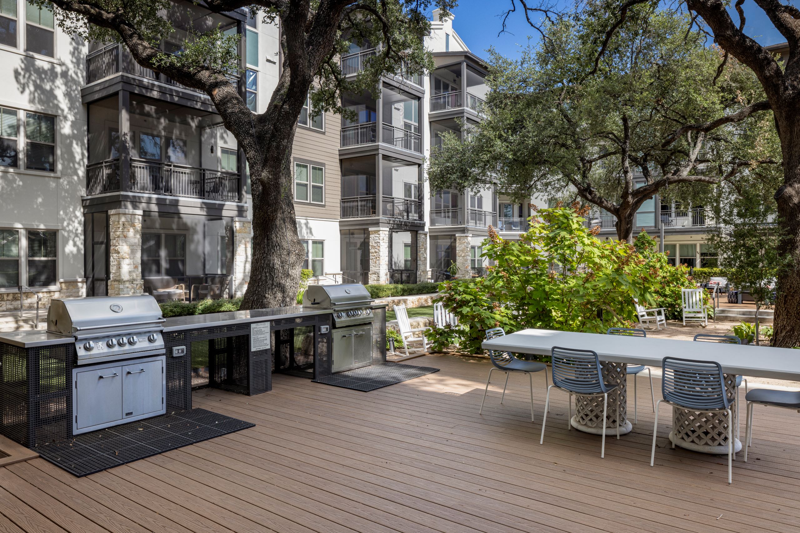 Two stainless steel grills and a long white dining table with six chairs surrounded by large trees and greenery in AMLI Lakeline courtyard