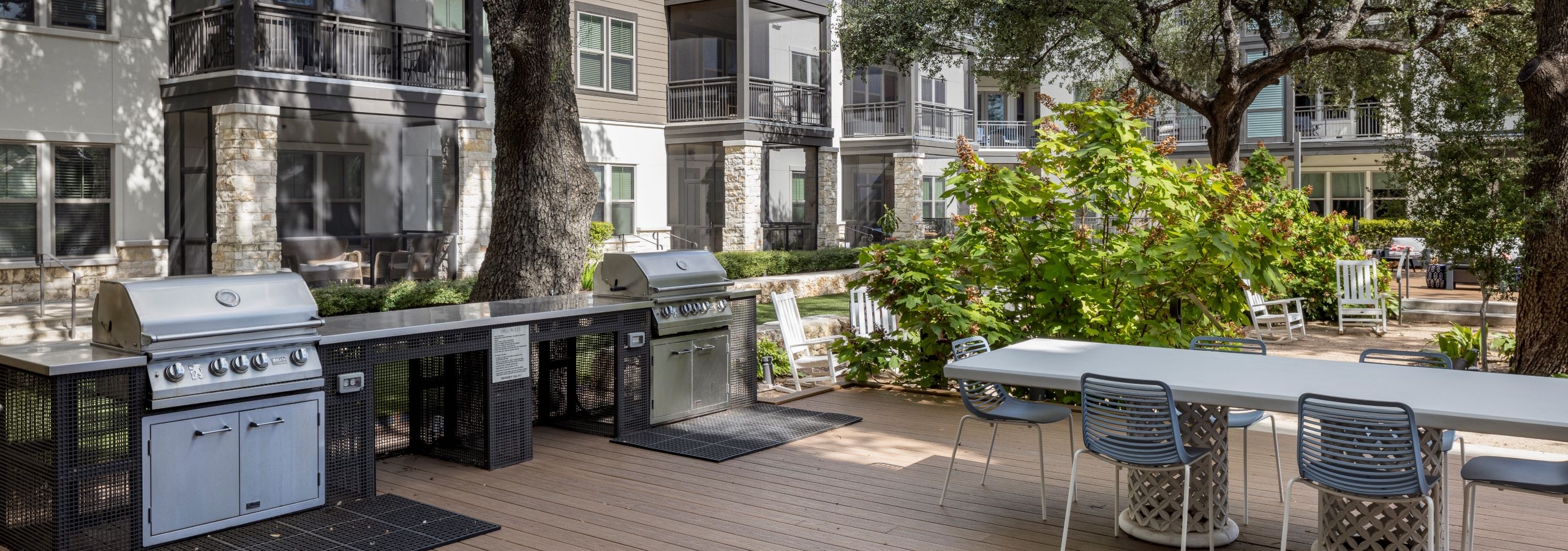 Two stainless steel grills and a long white dining table with six chairs surrounded by large trees and greenery in AMLI Lakeline courtyard