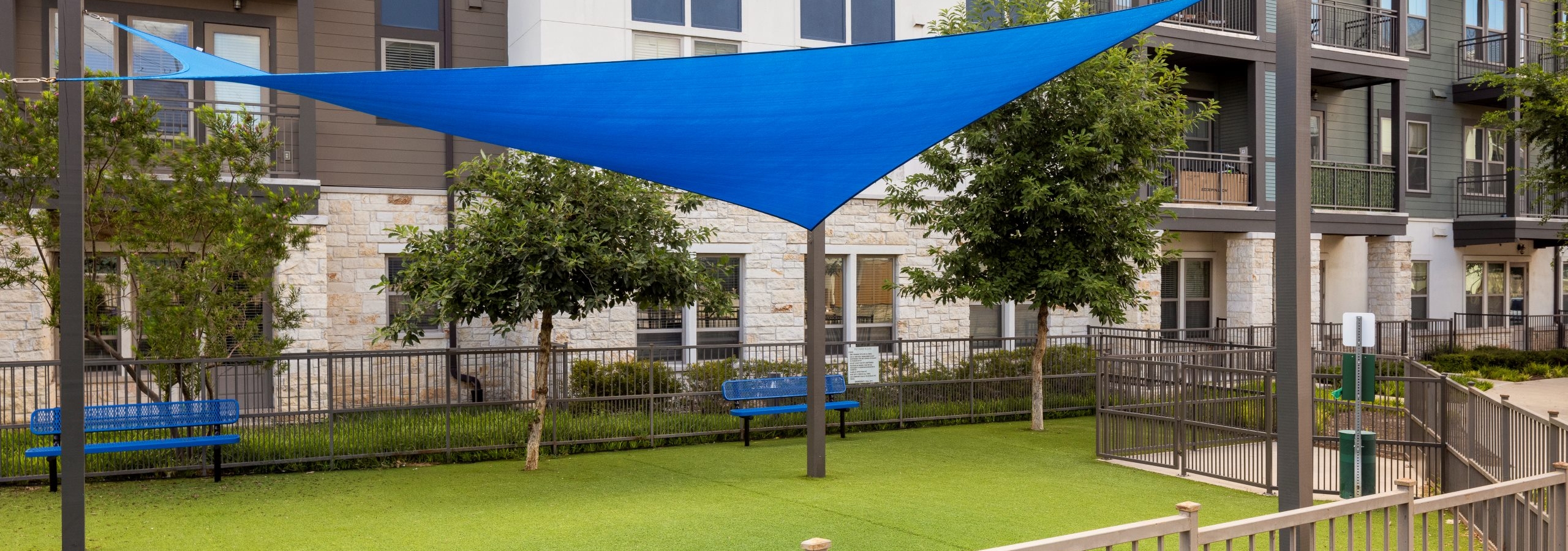 A fenced grassy pet park with bench seating shaded by a blue triangular cloth awning supported by metal poles at AMLI Lakeline apartments