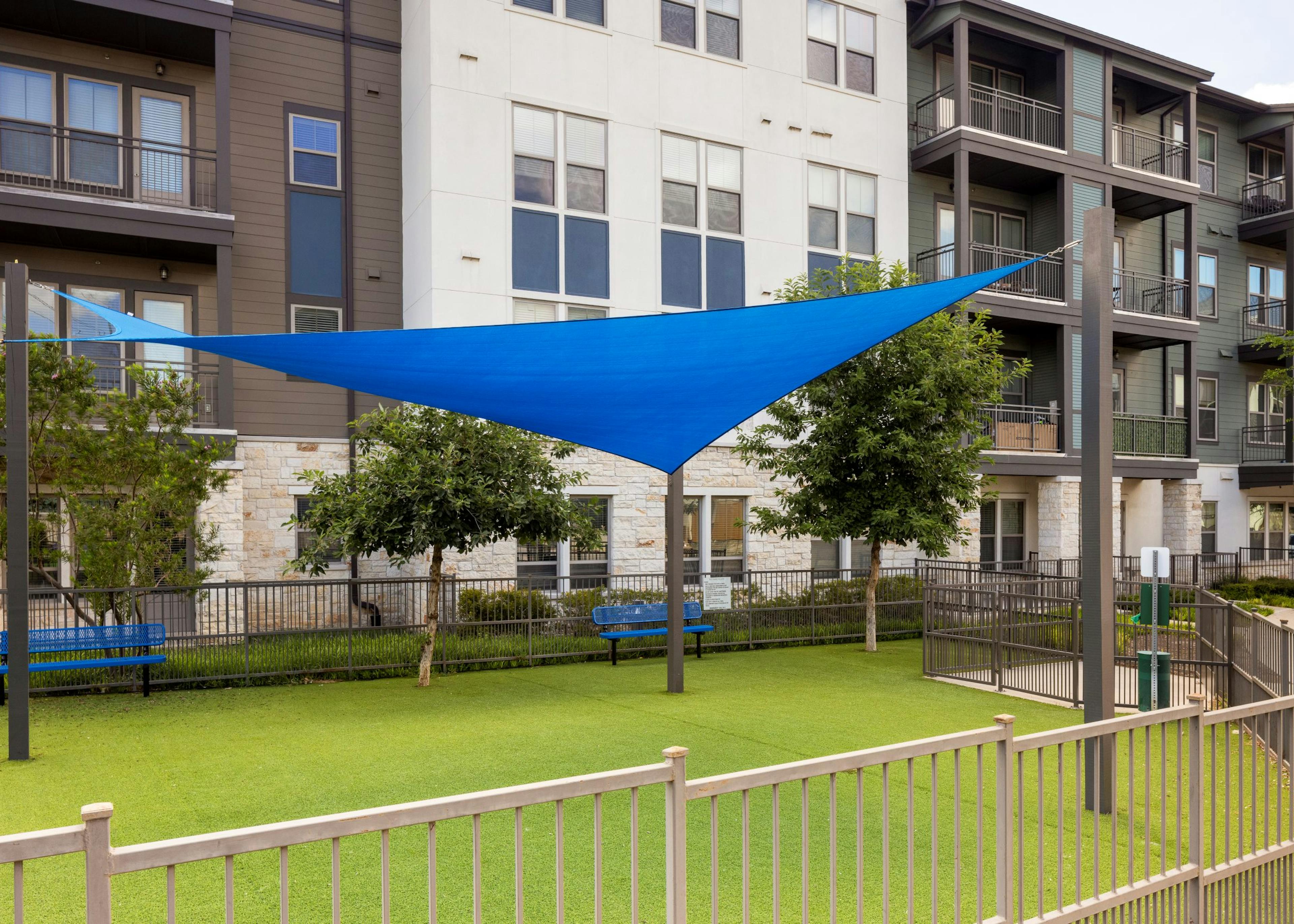 A fenced grassy pet park with bench seating shaded by a blue triangular cloth awning supported by metal poles at AMLI Lakeline apartments