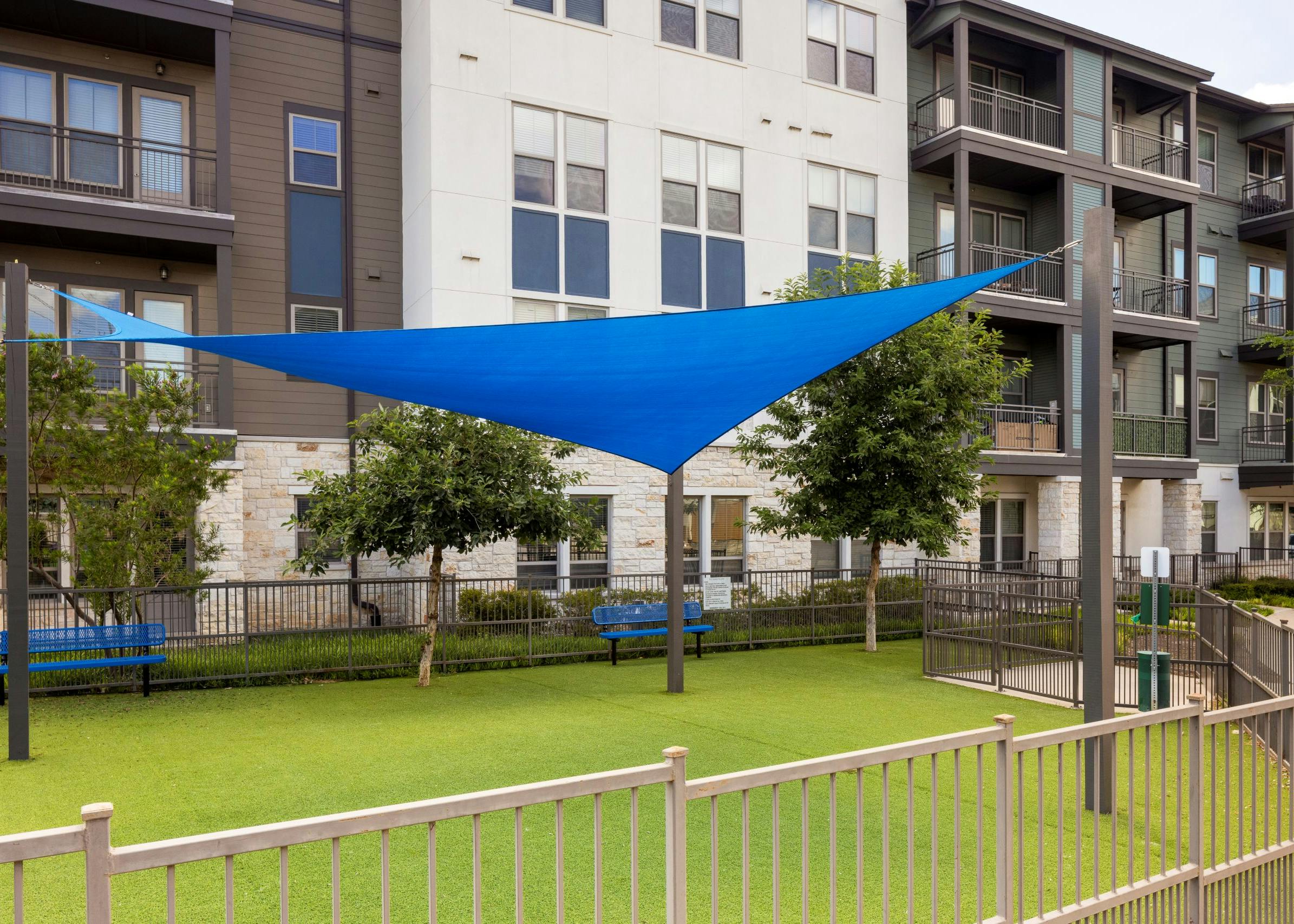 A fenced grassy pet park with bench seating shaded by a blue triangular cloth awning supported by metal poles at AMLI Lakeline apartments