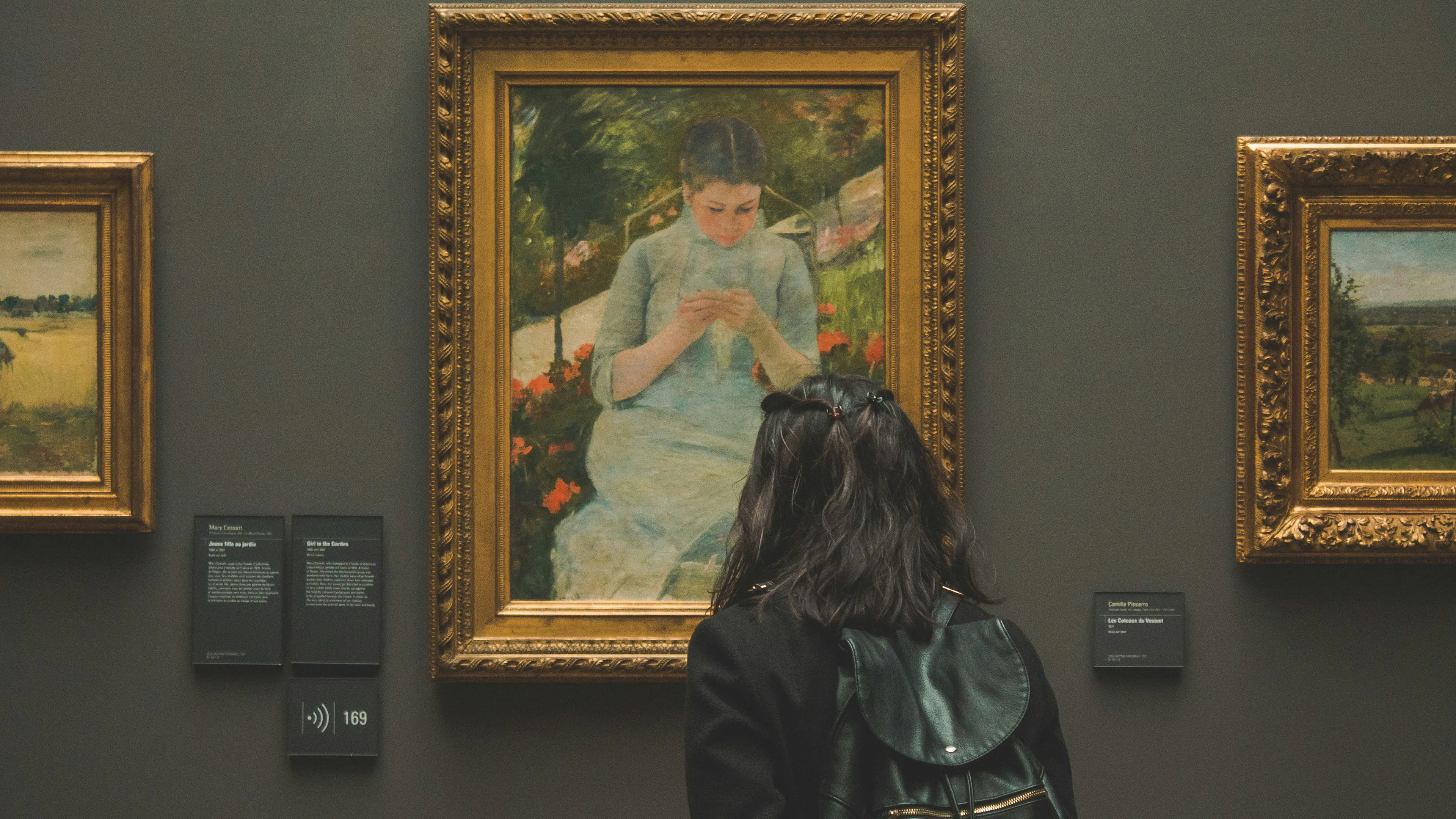 A woman facing away toward a large impressionist painting of a young girl sewing in a chair