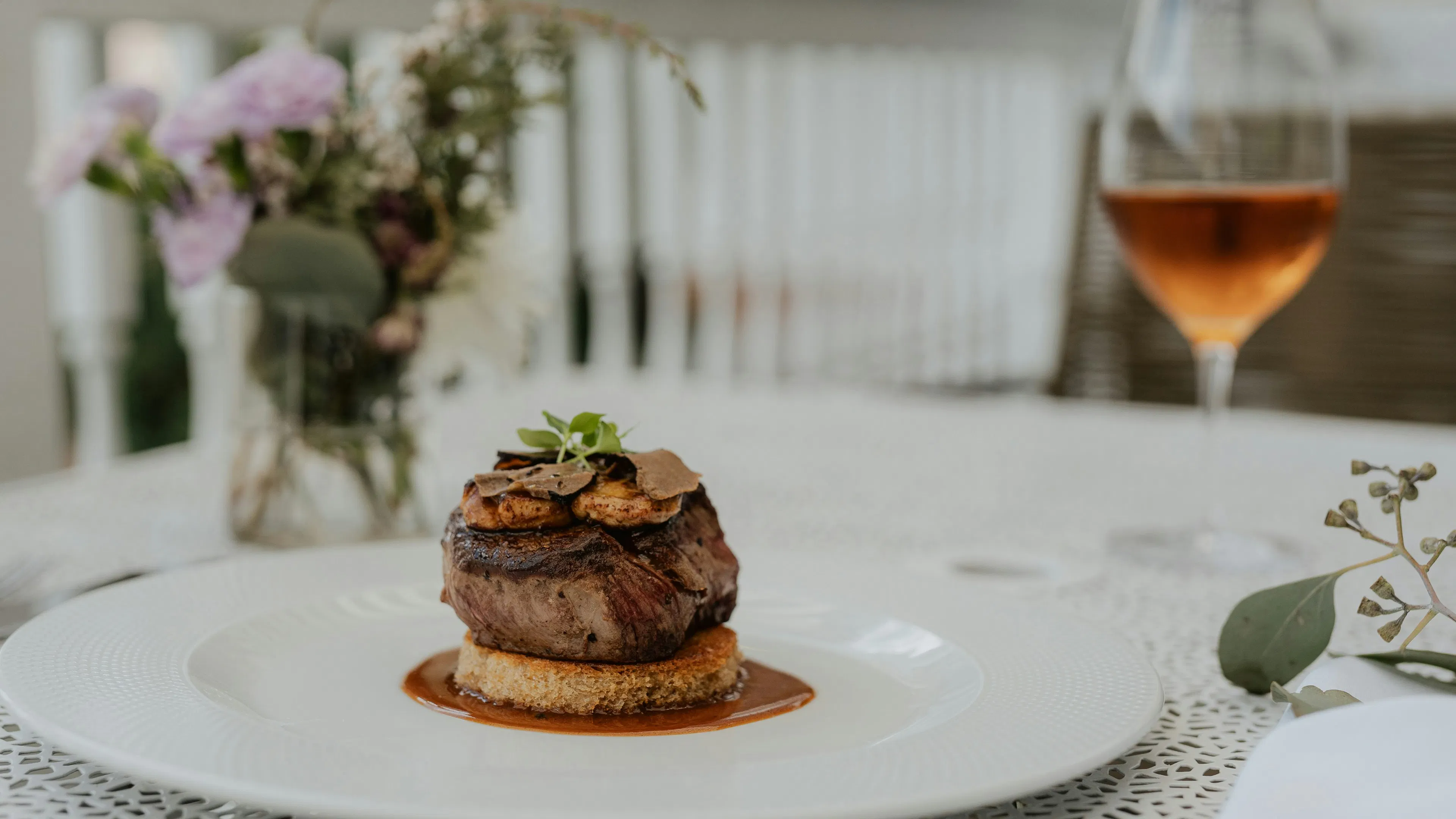 A small filet mignon with a red sauce on a white plate next to a glass of rose wine
