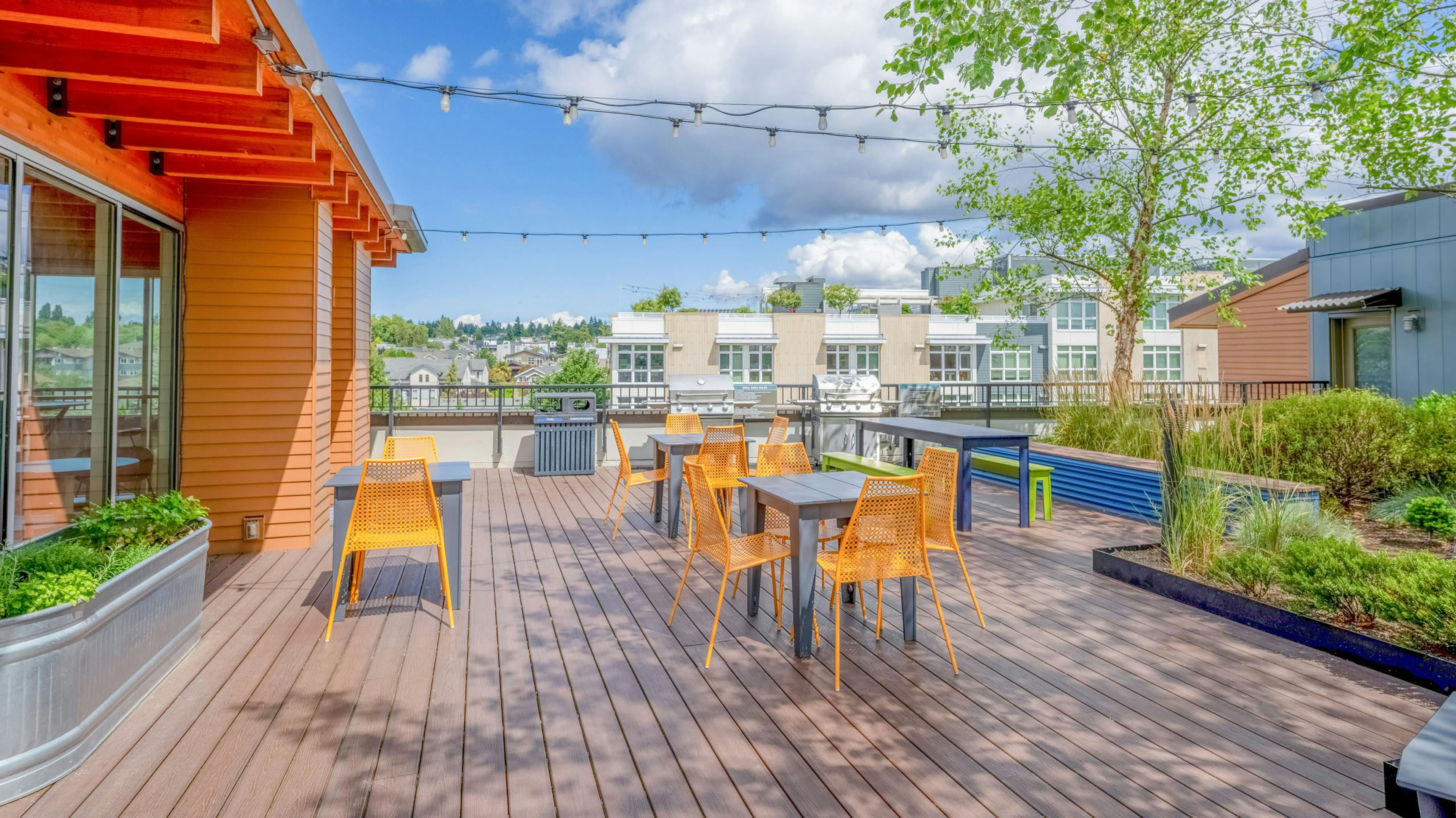 Rooftop at AMLI Mark24 featuring orange chairs around tables, string lights overhead, and stainless grill set on wooden decking.