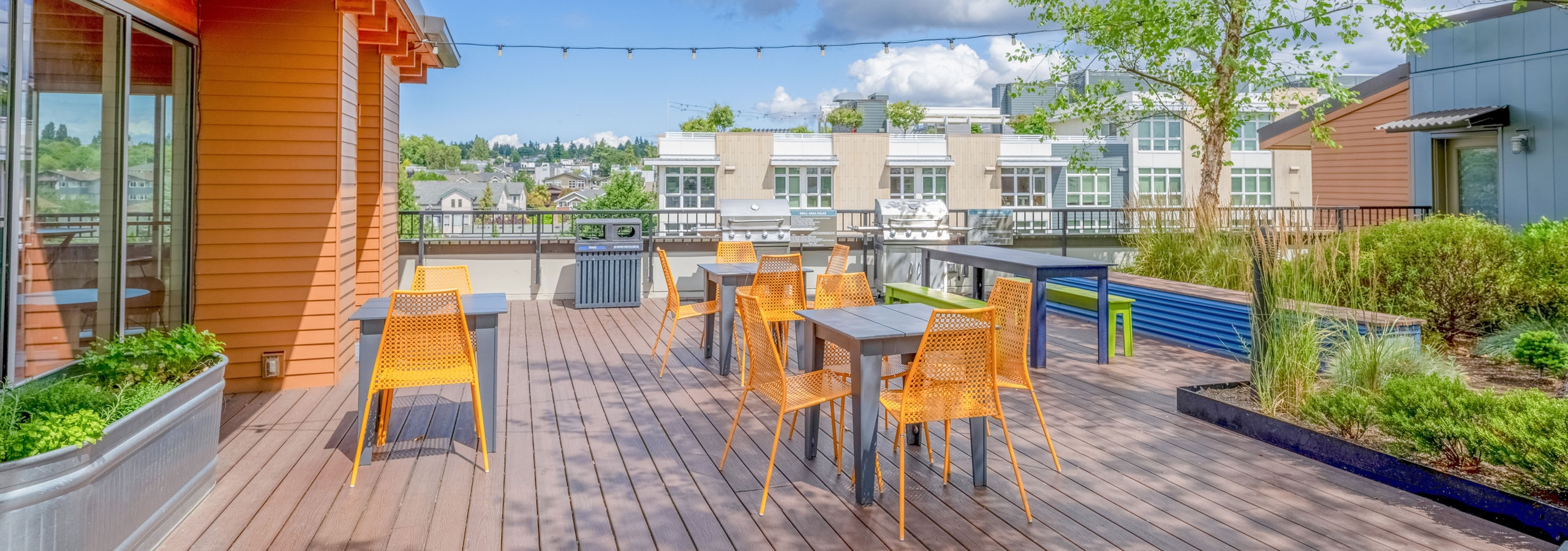 Rooftop at AMLI Mark24 featuring orange chairs around tables, string lights overhead, and stainless grill set on wooden decking.
