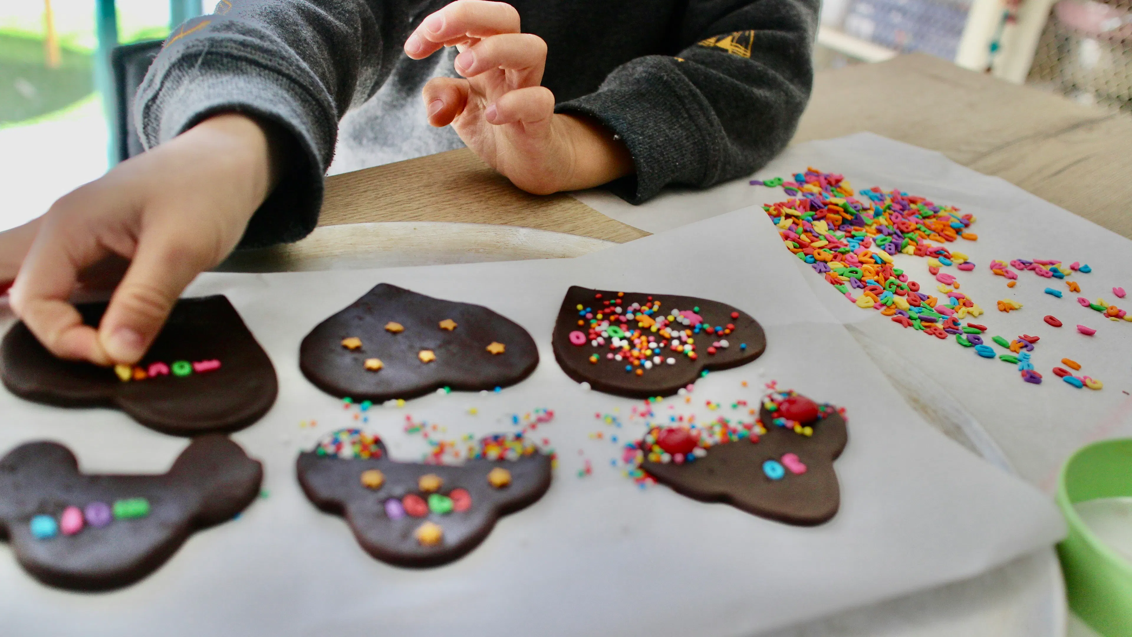 A kid putting sprinkles onto some chocolate cookies on a baking sheet
