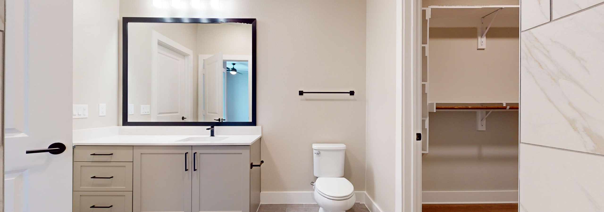 A spacious, modern bathroom featuring a white toilet, gray vanity and mirror with view into walk-in closet at  AMLI North Park apartments