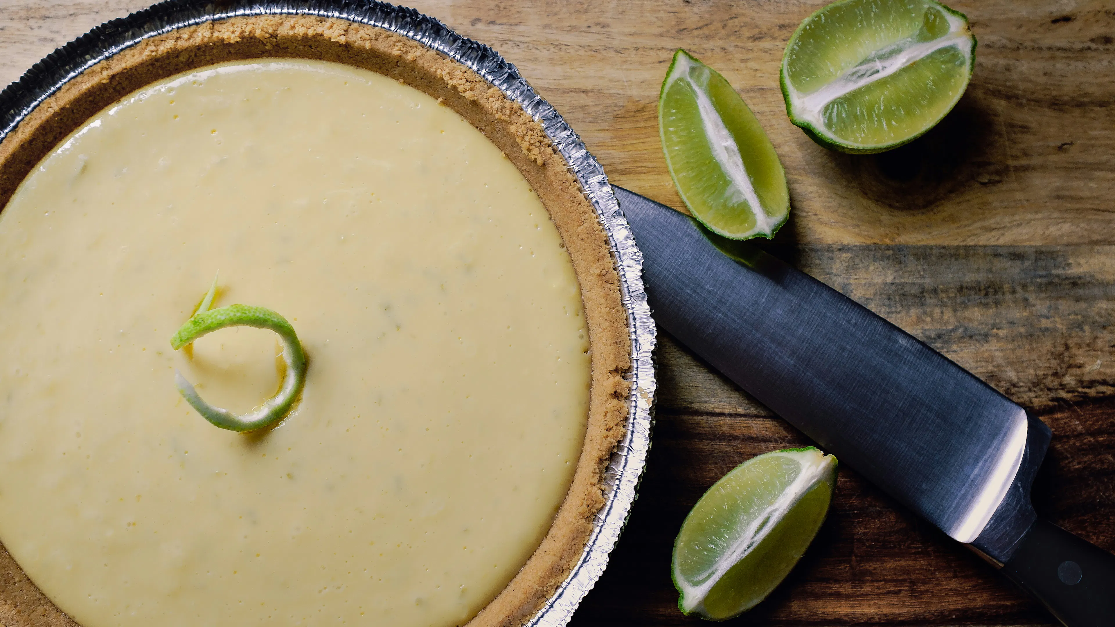 A key lime pie garnished with a twist of lime peel on a wooden table alongside some sliced limes
