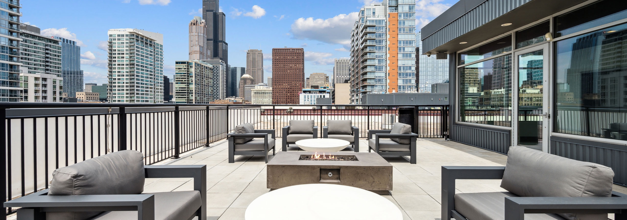 Rooftop lounge at AMLI Lofts apartments featuring gray chairs and circular white tables around a square fire pit overlooking city skyline.
