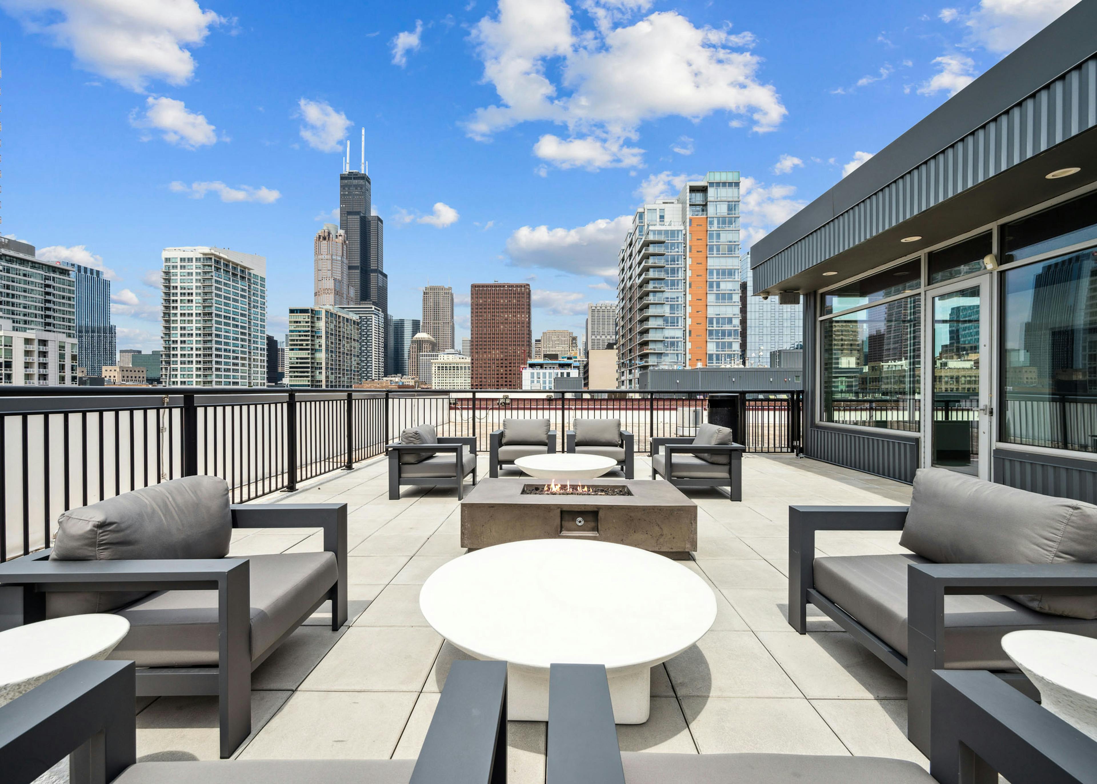 Rooftop lounge at AMLI Lofts apartments featuring gray chairs and circular white tables around a square fire pit overlooking city skyline.