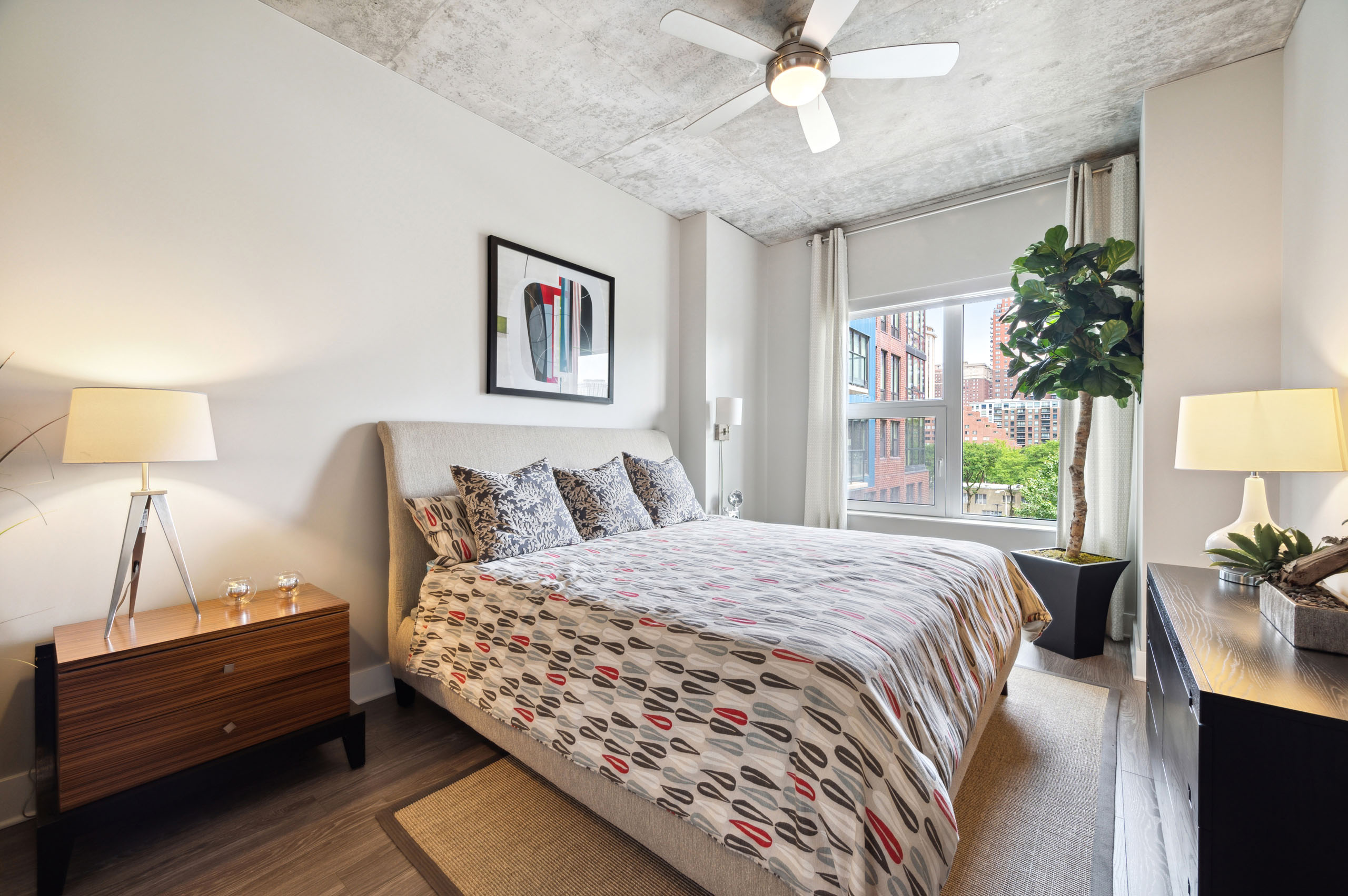 AMLI Lofts bedroom featuring a bed with a patterned duvet and two nightstands with lamps on either side with a large window with a city view.