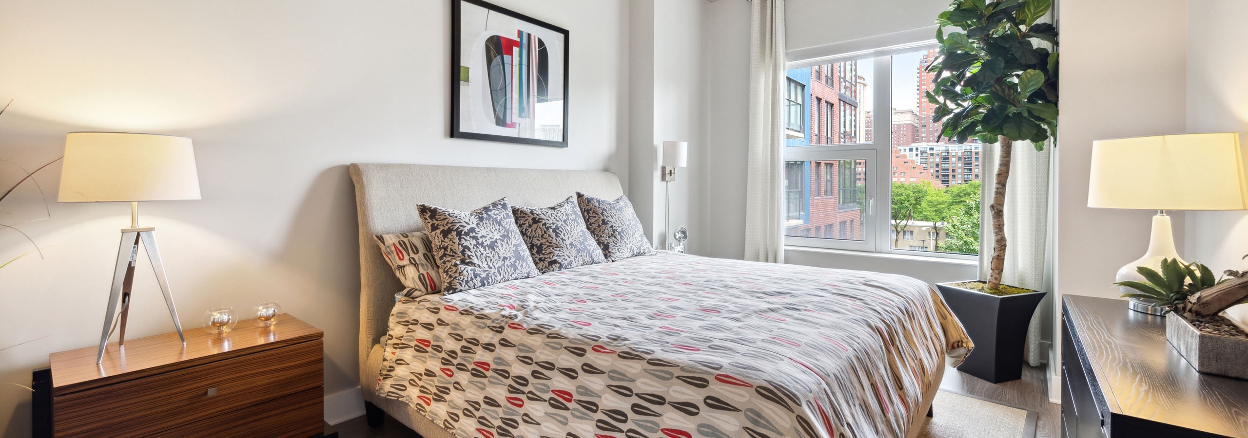 AMLI Lofts bedroom featuring a bed with a patterned duvet and two nightstands with lamps on either side with a large window with a city view.