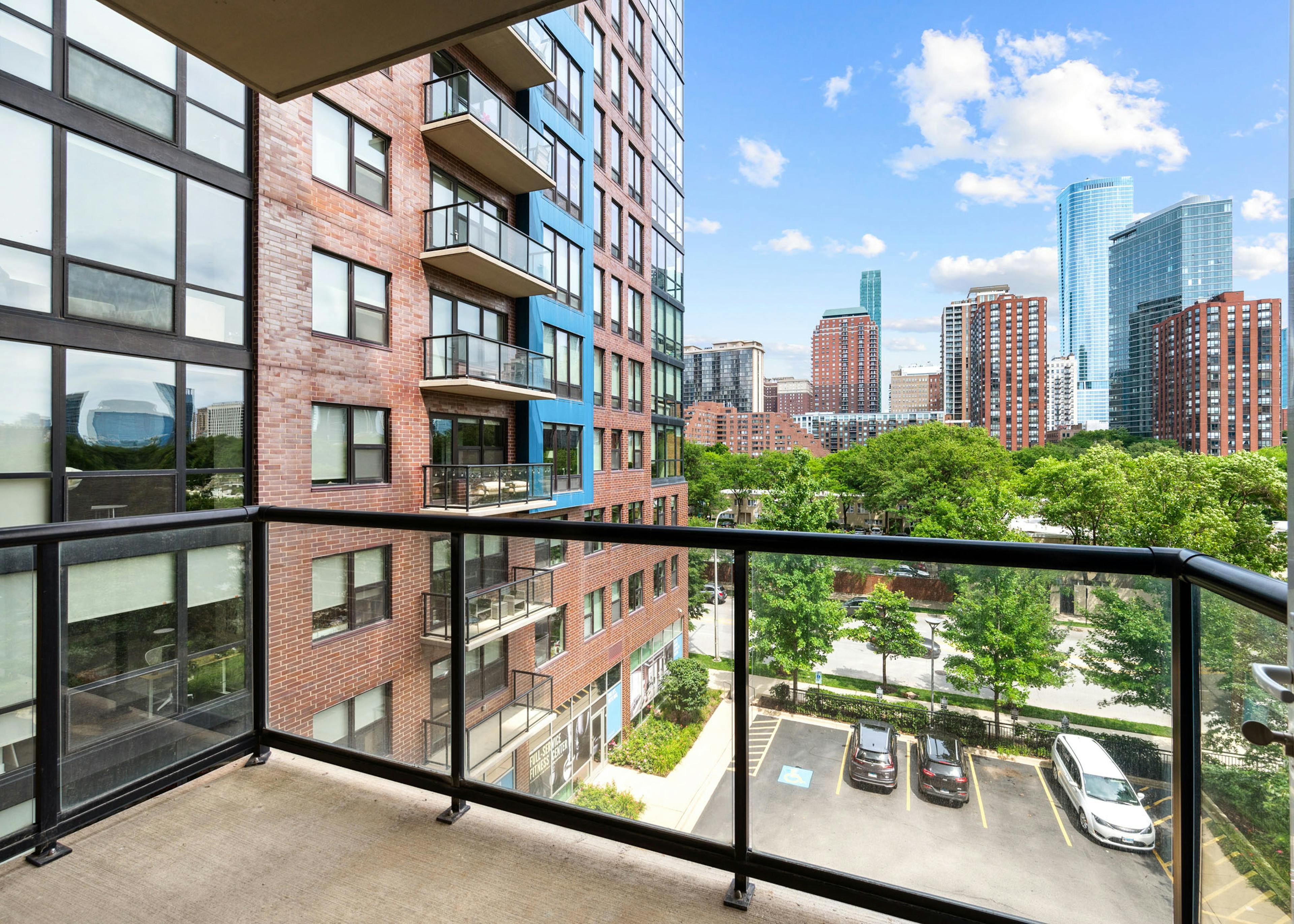 AMLI Lofts apartment balcony view overlooking a cityscape under a blue sky and the building’s parking lot and a tree line street.