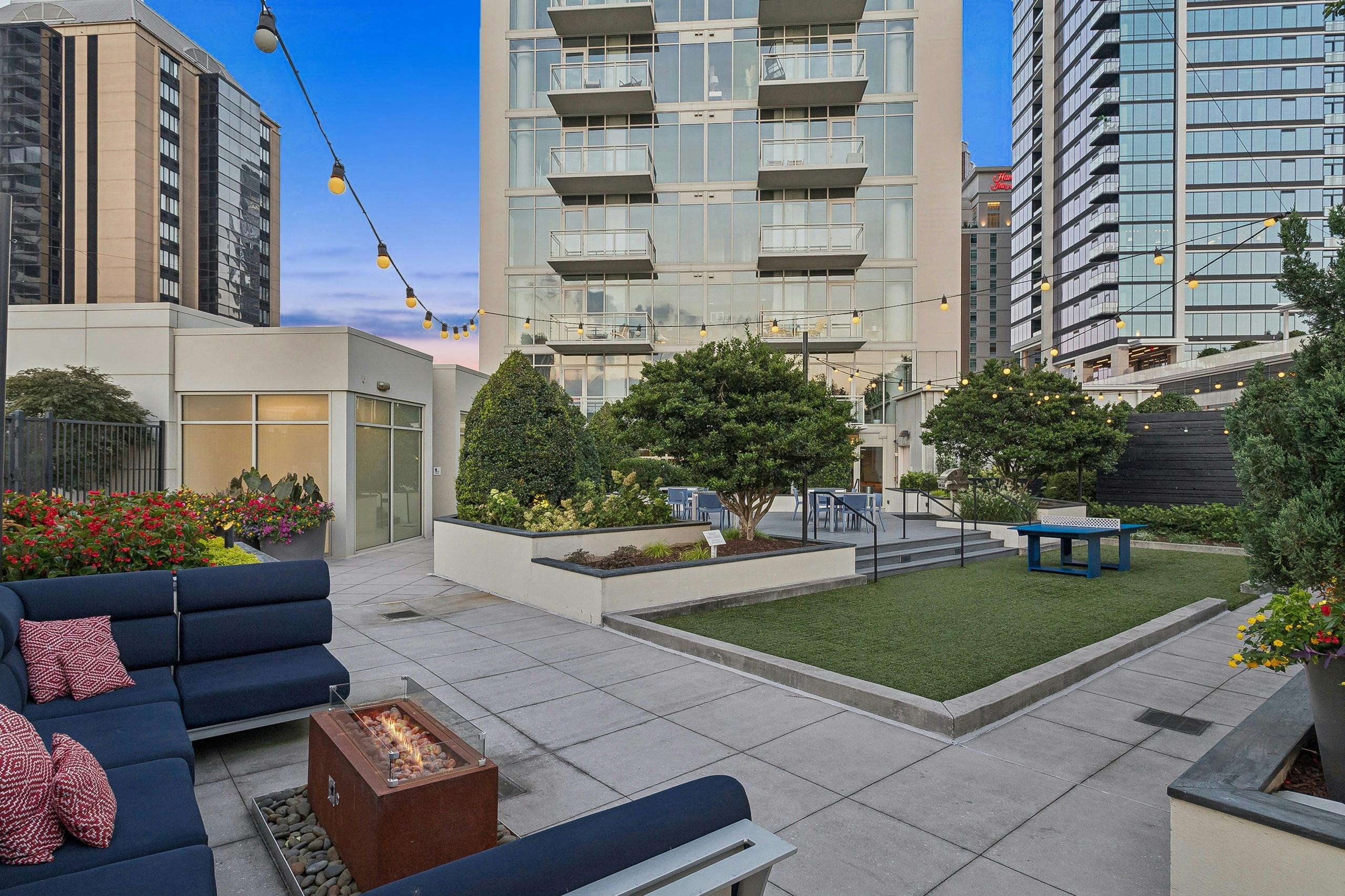 AMLI Arts Center rooftop patio with a fire pit and navy couch and turf area with ping pong table surrounded by trees and flowers