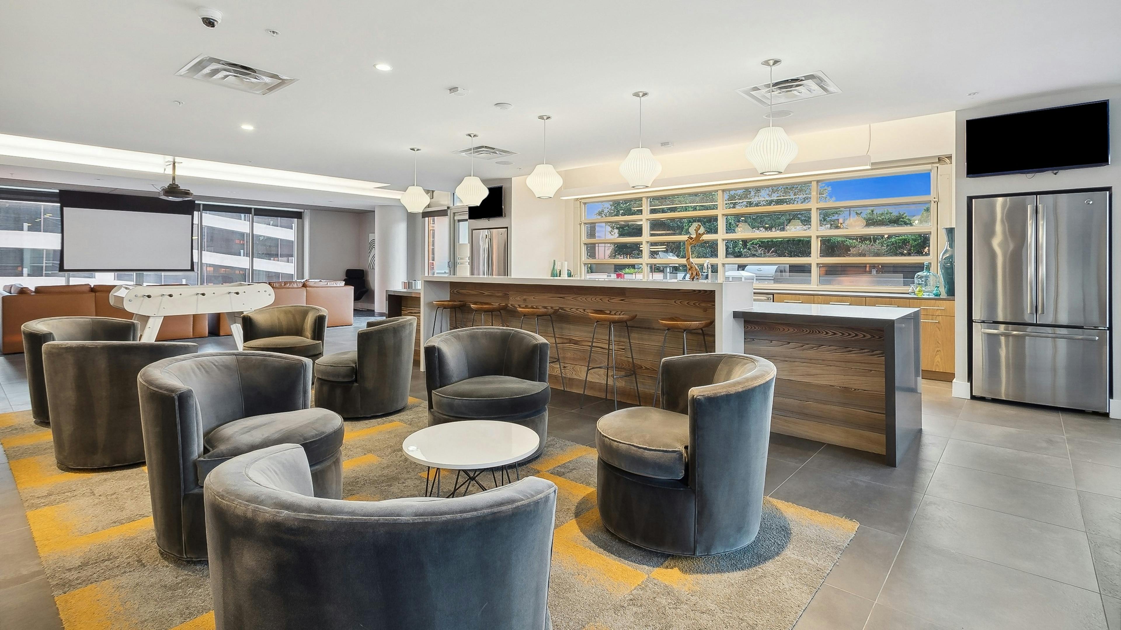 Clubroom at AMLI Arts Center with gray velvet chairs around tables and a long kitchen counter with wooden stools, foosball and movie screen