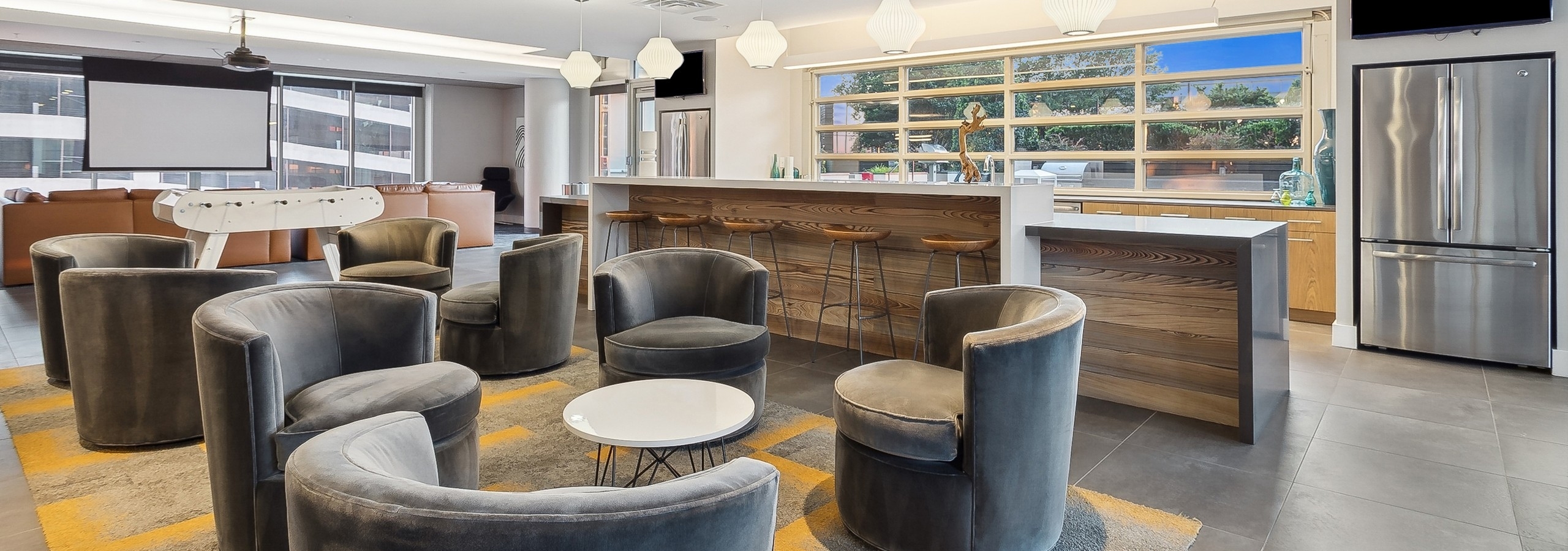 Clubroom at AMLI Arts Center with gray velvet chairs around tables and a long kitchen counter with wooden stools, foosball and movie screen