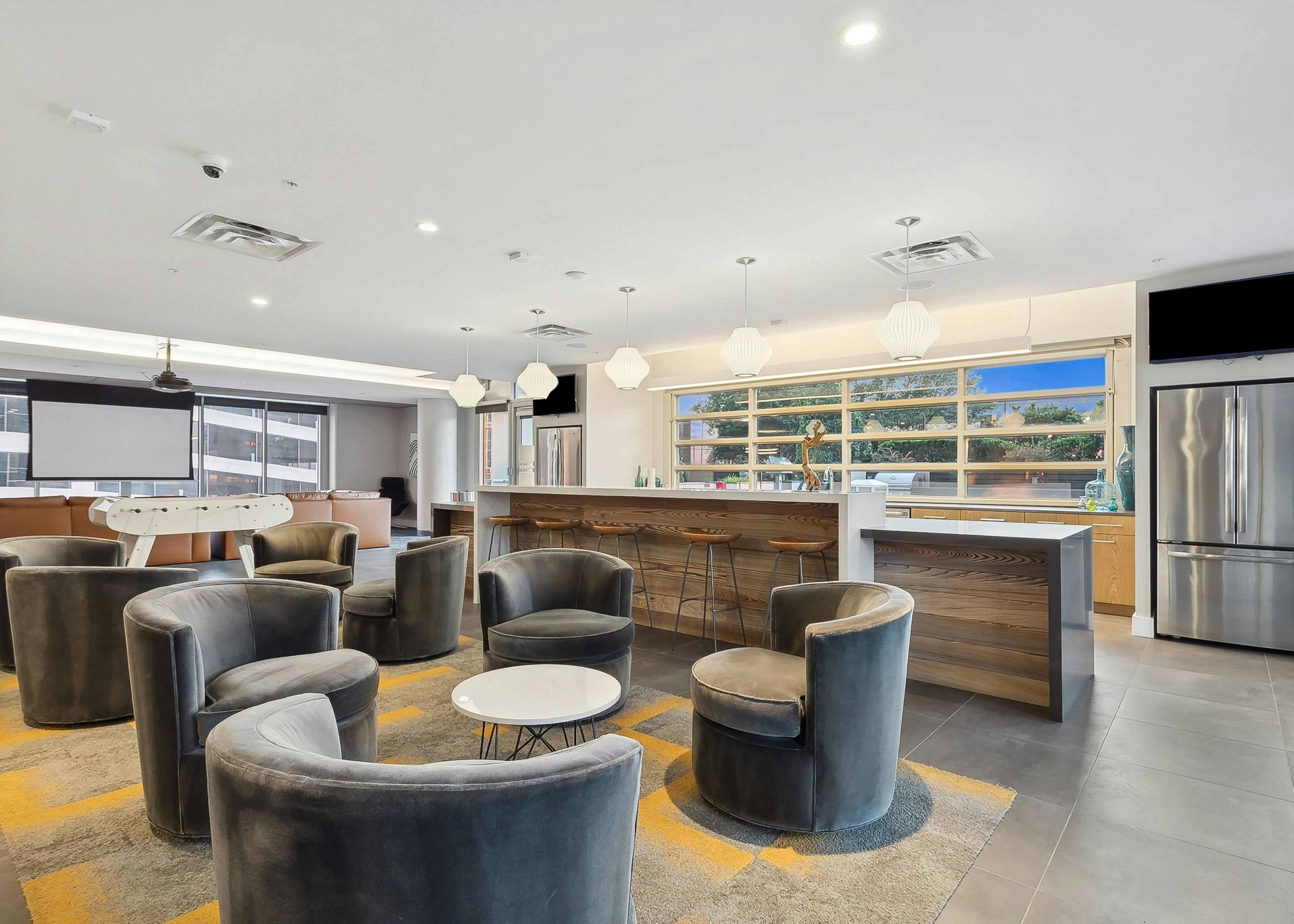 Clubroom at AMLI Arts Center with gray velvet chairs around tables and a long kitchen counter with wooden stools, foosball and movie screen