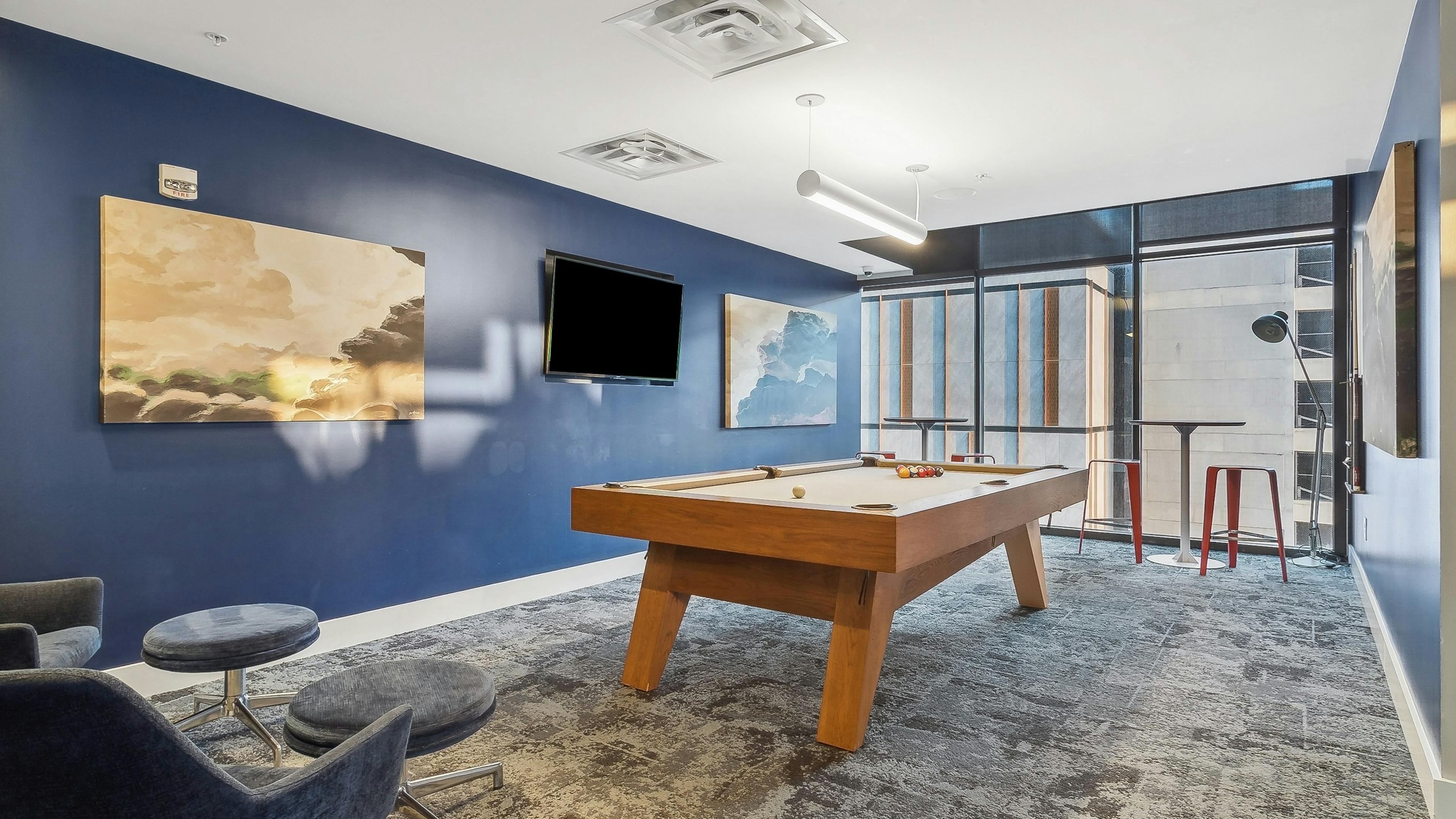 Game room at AMLI Arts Center with a pool table in the center with and abstract paintings and a wall mounted television on a blue wall