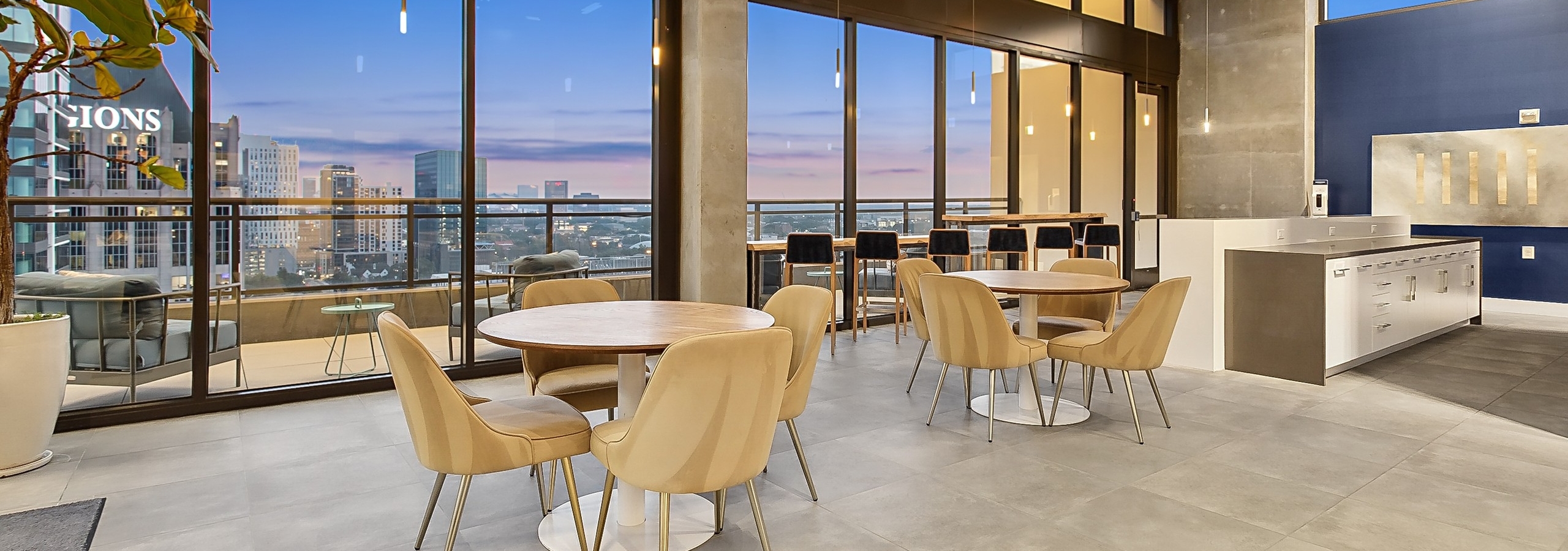 A chic lounge area at AMLI Arts Center with tables and chairs and large floor to ceiling windows showcasing a cityscape at sunset