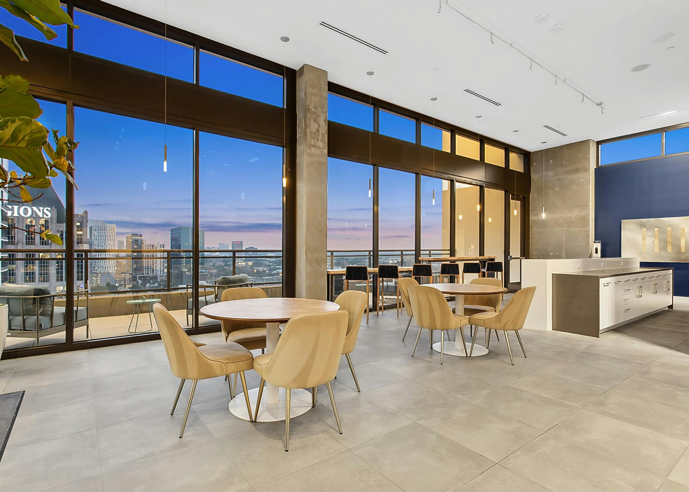 A chic lounge area at AMLI Arts Center with tables and chairs and large floor to ceiling windows showcasing a cityscape at sunset