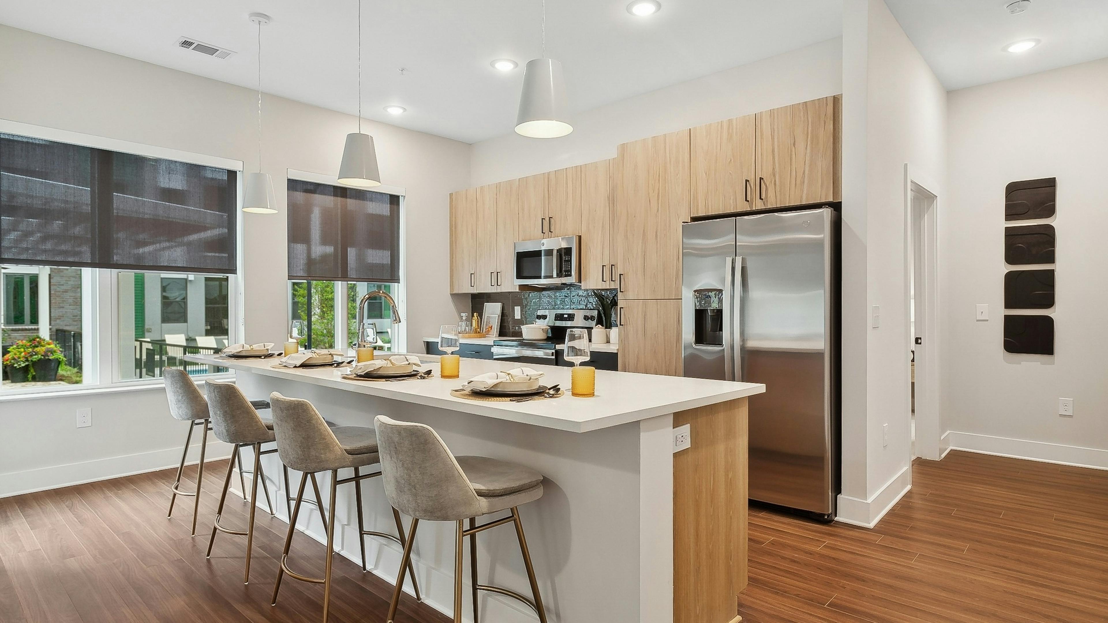 AMLI Flatiron kitchen with a long white island and four gray barstools and pendant lights and wood cabinets and stainless steel appliances