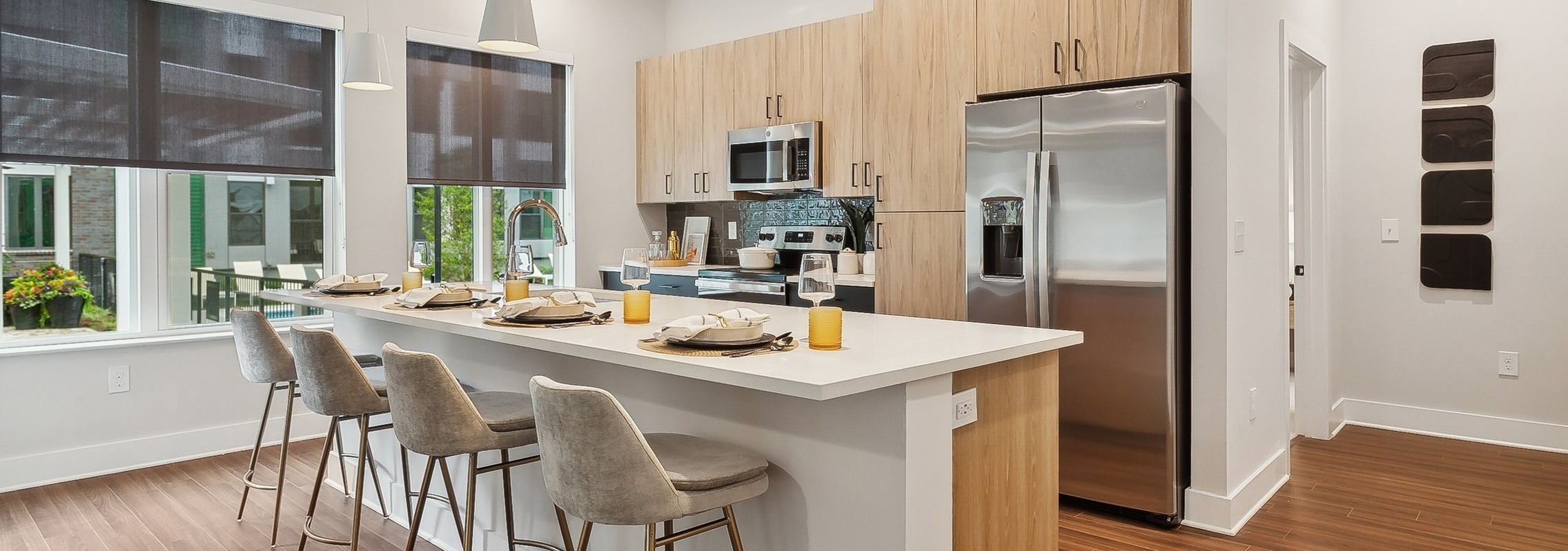 AMLI Flatiron kitchen with a long white island and four gray barstools and pendant lights and wood cabinets and stainless steel appliances