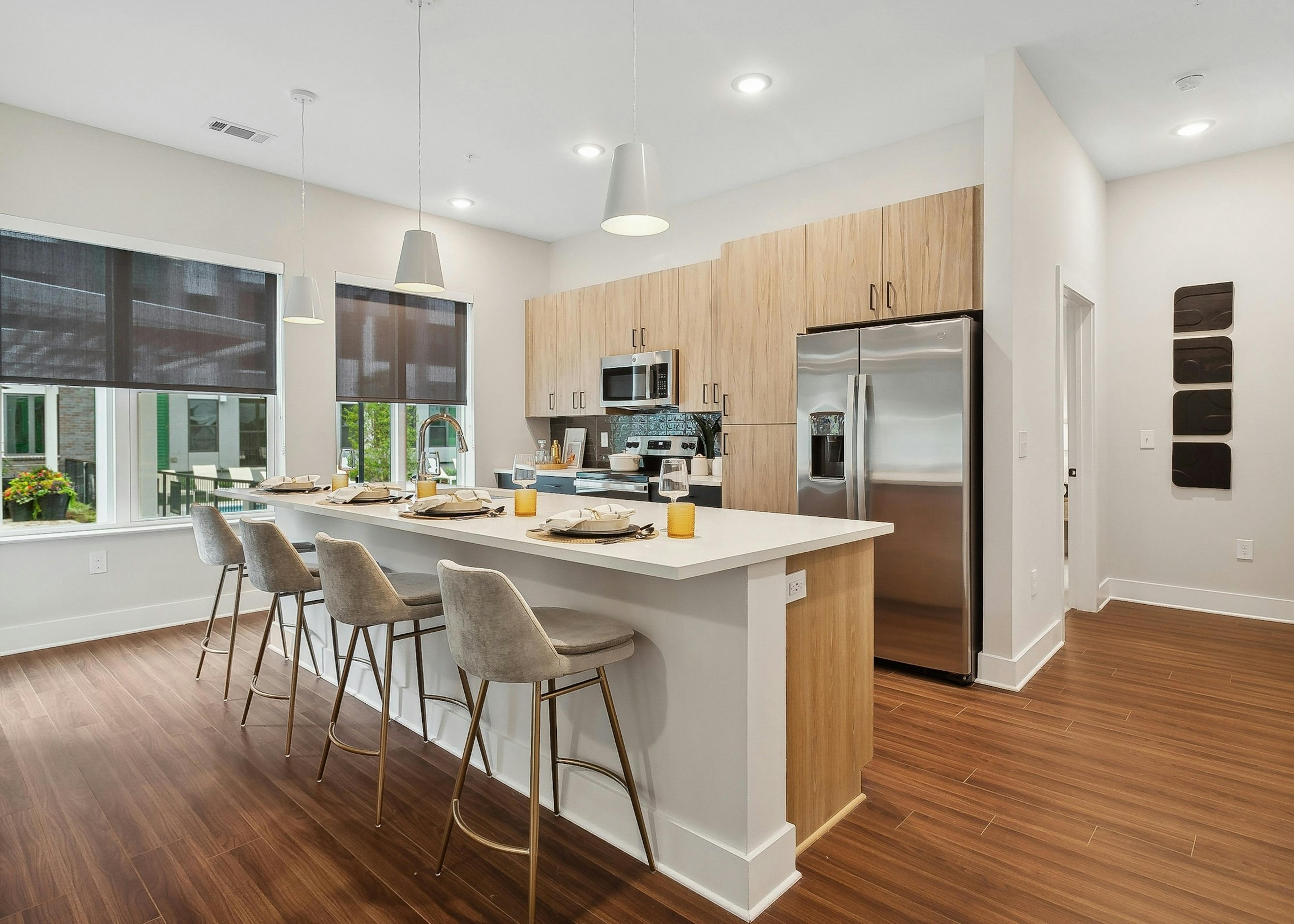 AMLI Flatiron kitchen with a long white island and four gray barstools and pendant lights and wood cabinets and stainless steel appliances