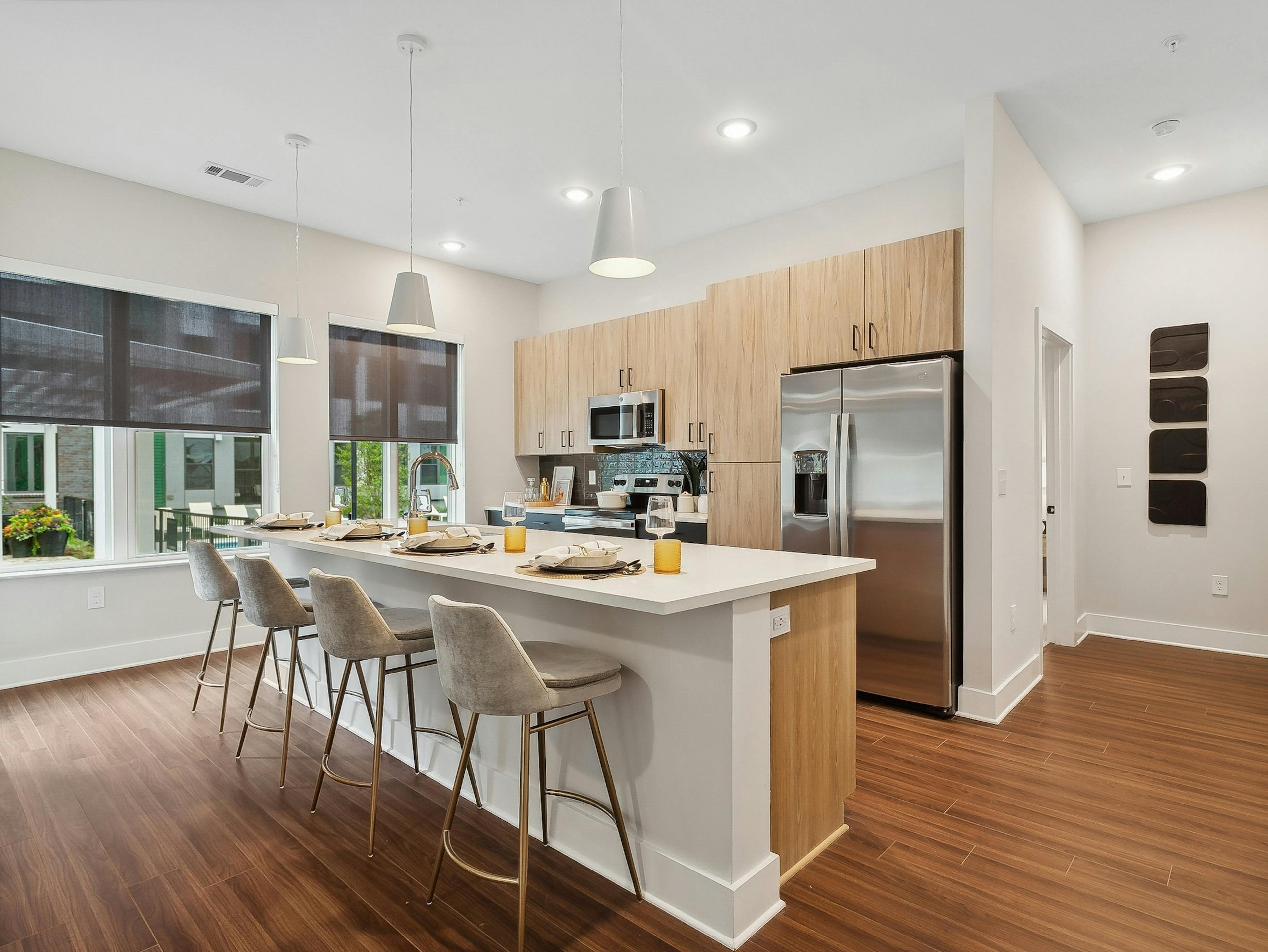 AMLI Flatiron kitchen with a long white island and four gray barstools and pendant lights and wood cabinets and stainless steel appliances
